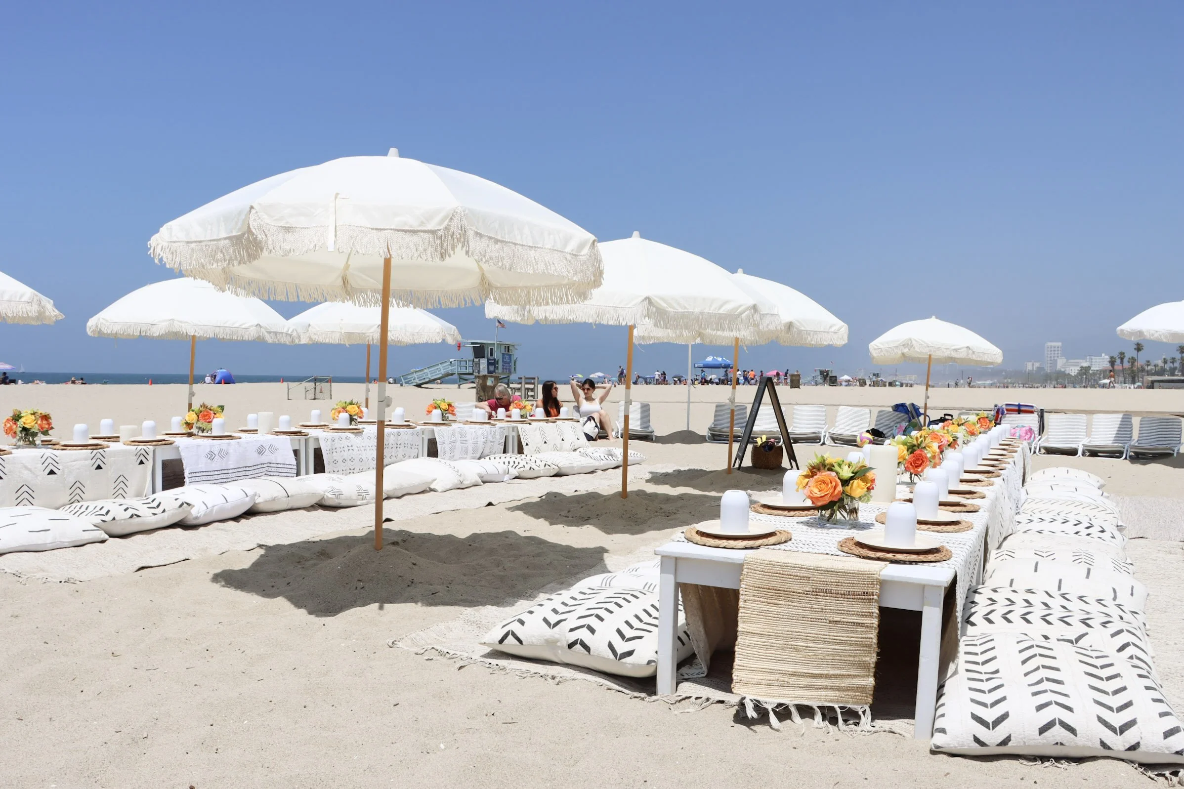 Beachside dining setup with white umbrellas, tables with floral centerpieces, plates, glasses, and cushions on sand, with people relaxing in the background and a clear blue sky.