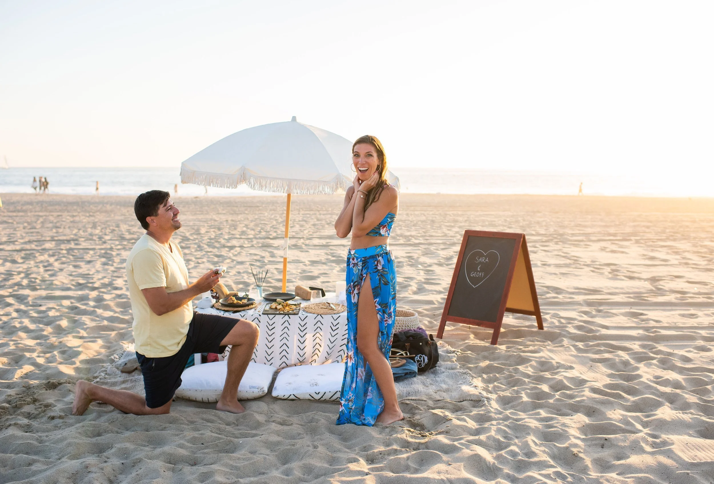 A couple enjoying a picnic on the beach during sunset, with a table set with food, an umbrella, and a chalkboard sign reading 'Sara & Goff'.
