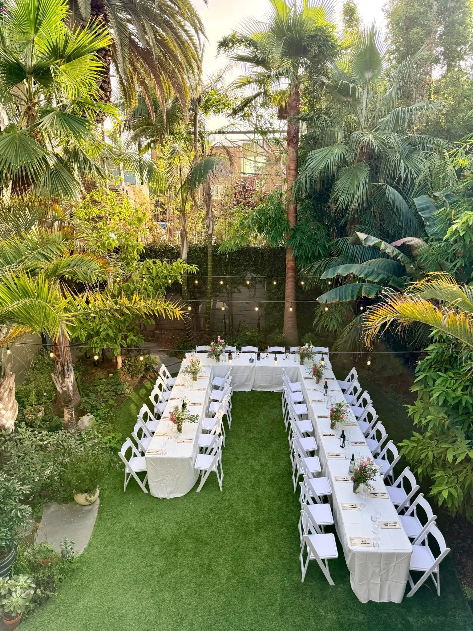 An outdoor dining setup in a lush, tropical garden with long white tables, white chairs, floral centerpieces, and string lights overhead.