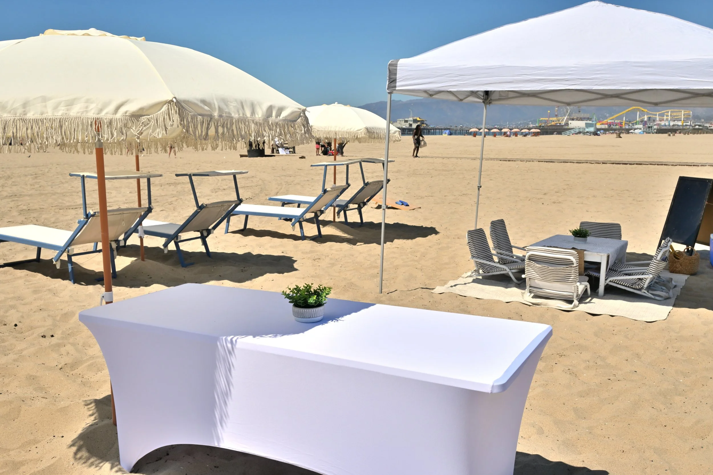 Beach scene with a shaded picnic area with a white table, striped chairs, coolers, and umbrellas. In the background, there are lounge chairs, umbrellas, and people on the sandy beach near the ocean, with a lifeguard tower and equipment visible on the right.
