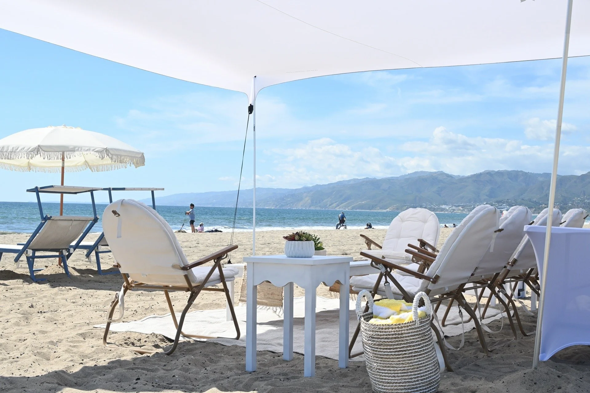 Beach scene with a shaded picnic area with a white table, striped chairs, coolers, and umbrellas. In the background, there are lounge chairs, umbrellas, and people on the sandy beach near the ocean, with a lifeguard tower and equipment visible on the right.