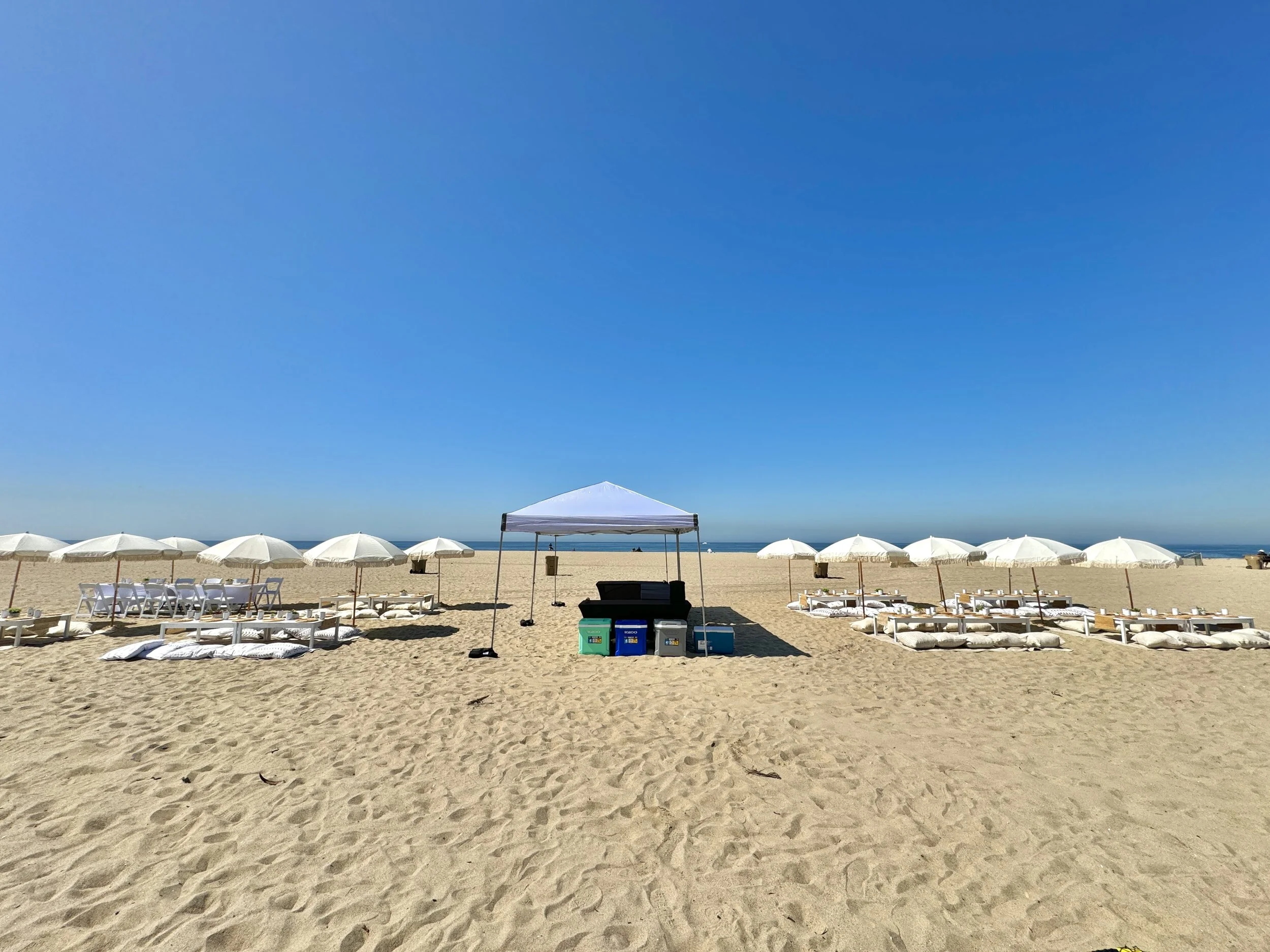 Empty beach with white umbrellas, lounge chairs, and cushions arranged along the sandy shore, with a white canopy tent in the center and the ocean in the background under a clear blue sky.