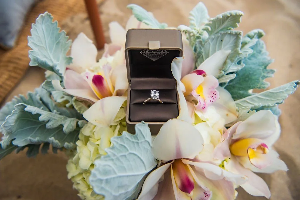 A diamond engagement ring in an open gray box, placed on a bouquet of white and pink orchids with large green leaves.