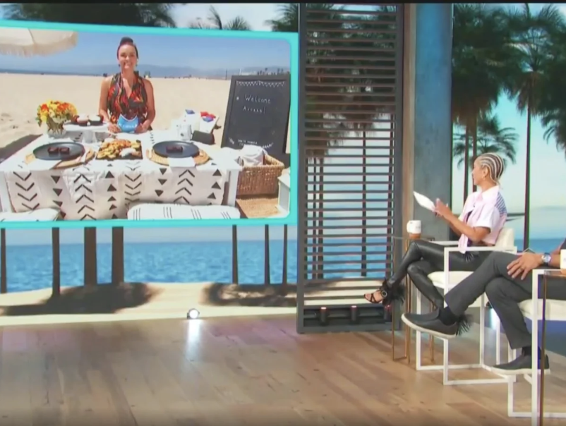 Television studio scene with two women sitting on white chairs, one holding a tablet and the other with a glass of drink, watching a large screen showing a woman at a beachside dining table with food and a welcome sign.