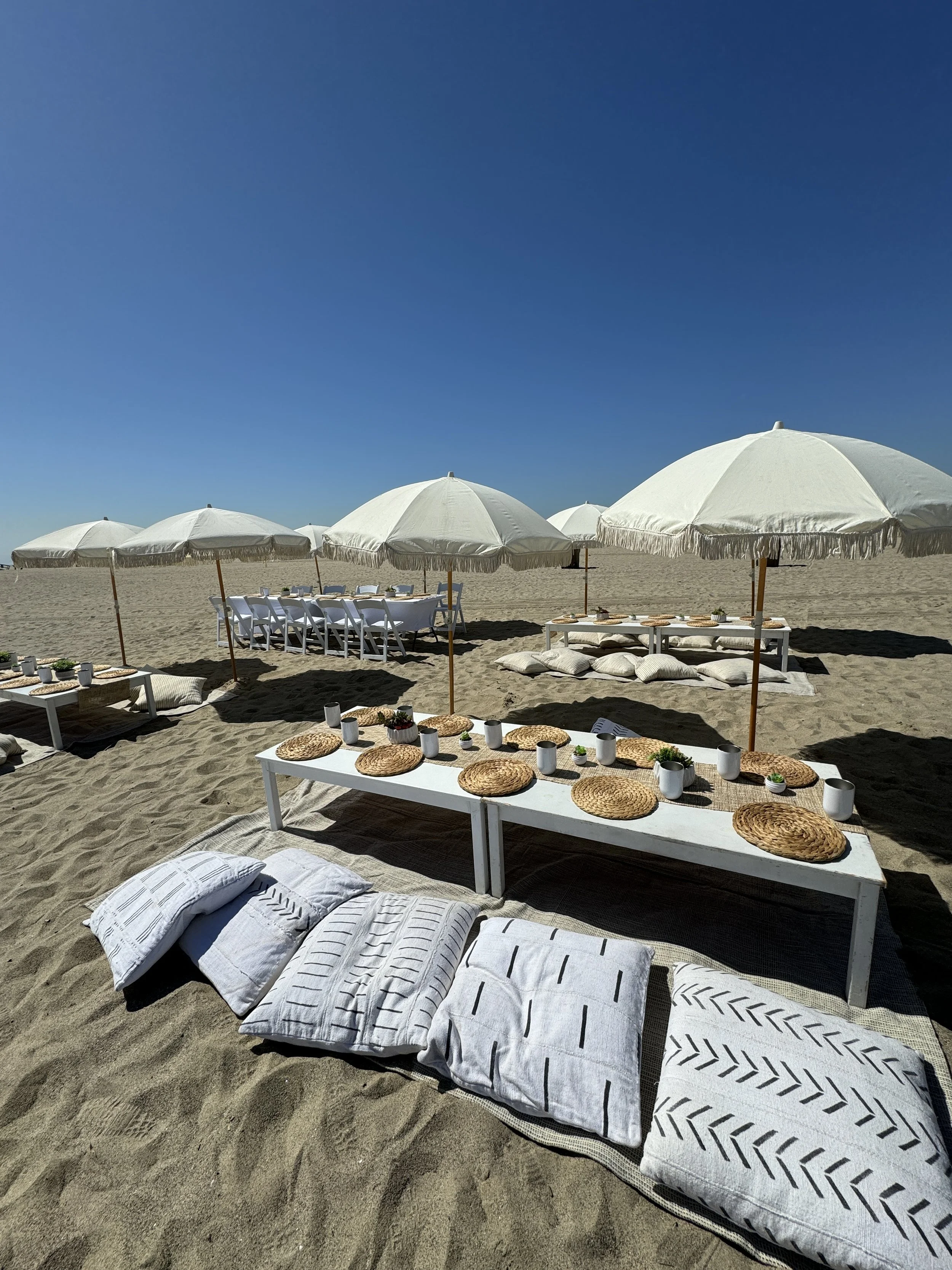 Beach setup with white umbrellas, low tables with cushions, woven placemats, and cups, on sand with a clear blue sky.