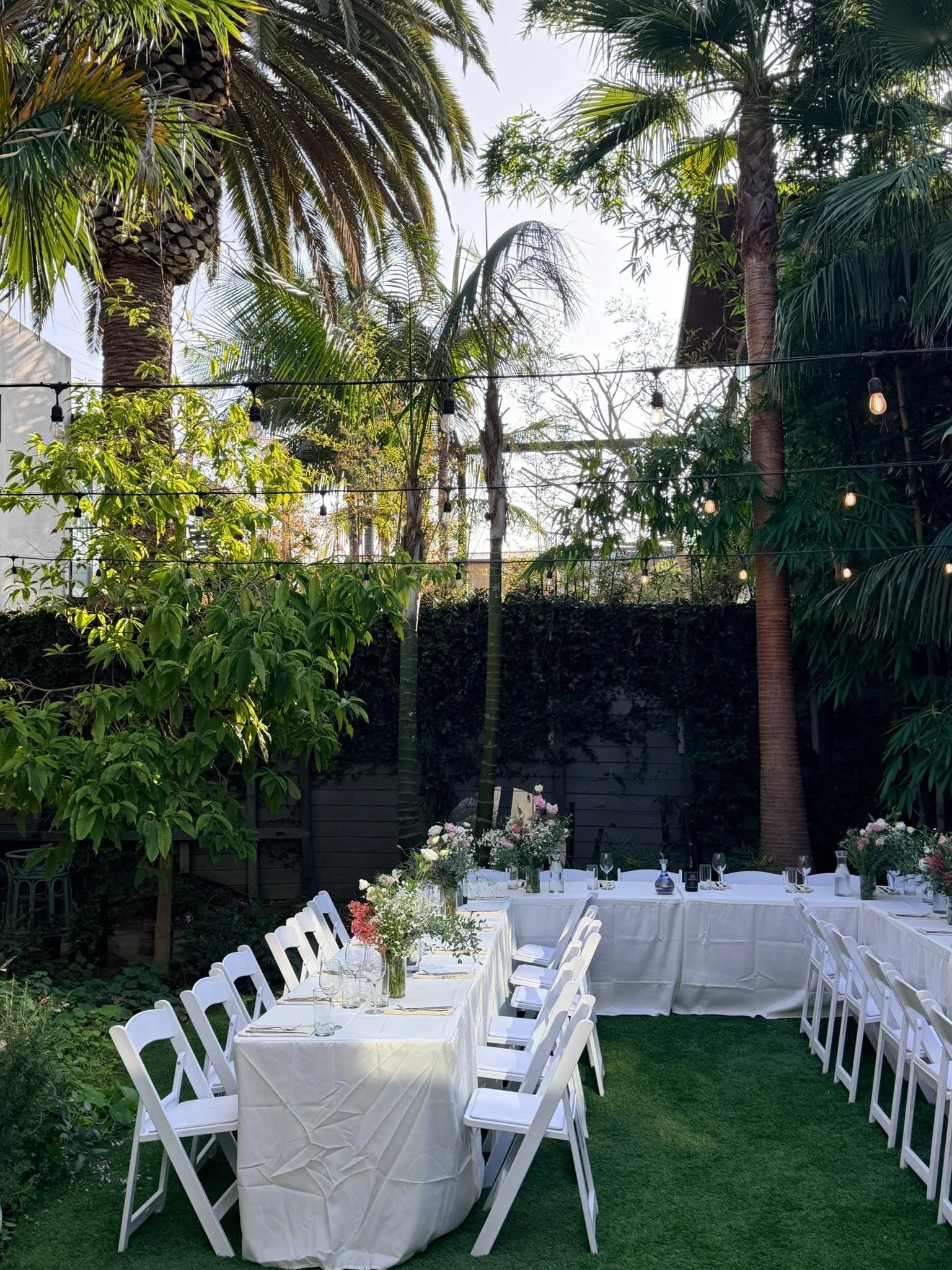 Outdoor dining setup with white tablecloths, surrounded by white chairs, decorated with floral centerpieces, under string lights, amidst lush green trees and palm trees in a garden.