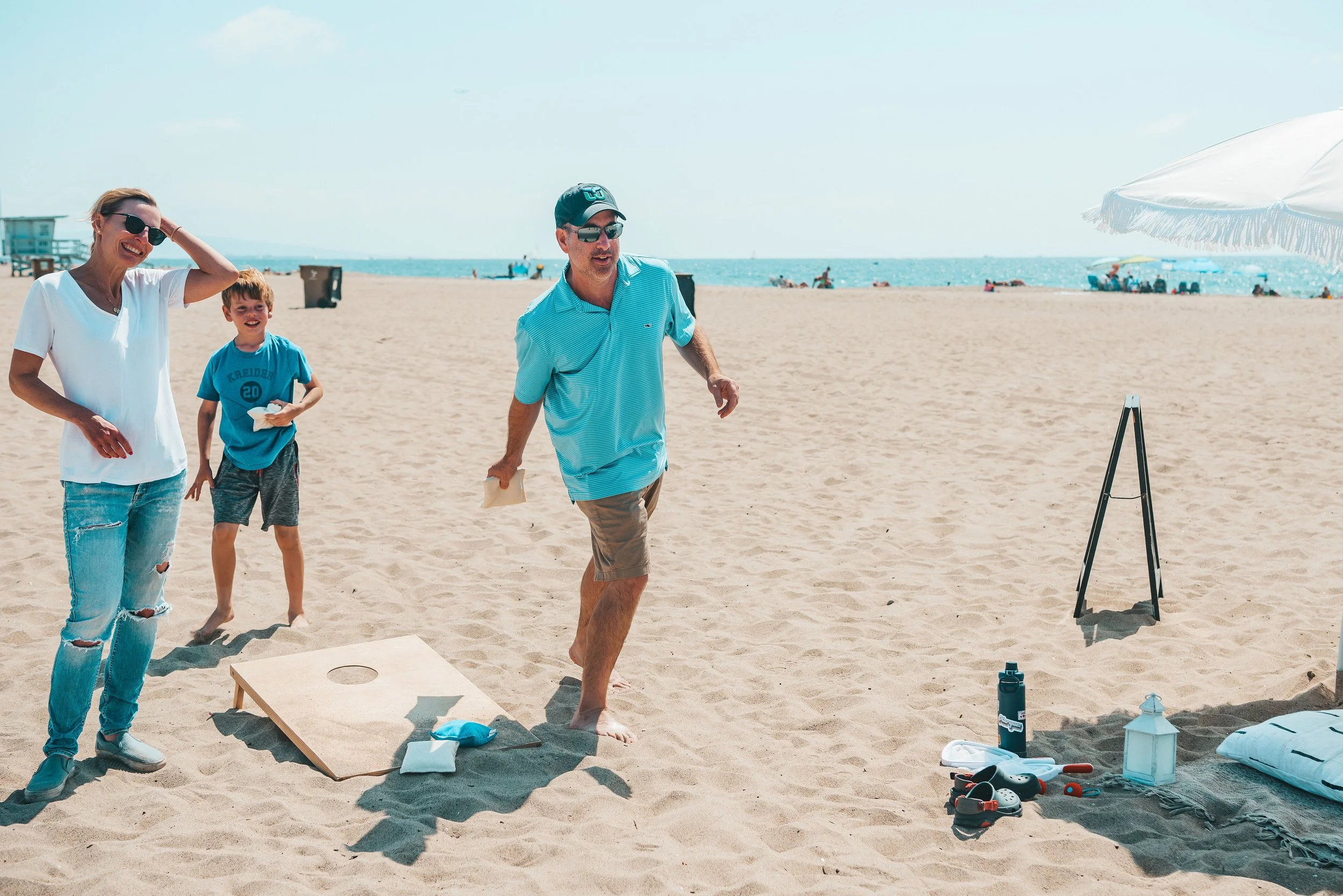 Family playing cornhole on the beach with umbrellas and people in the background.