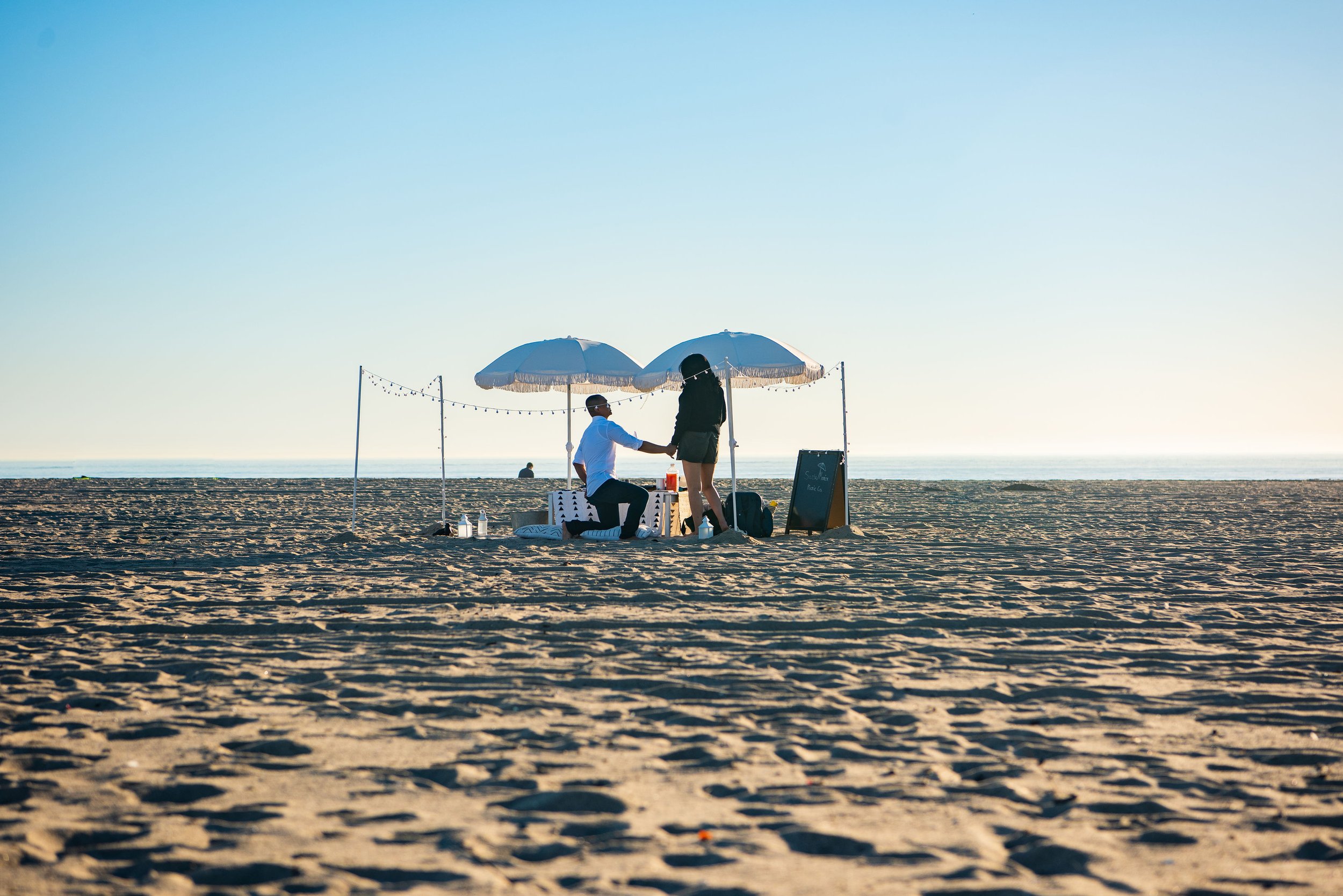 Couple on the beach under umbrellas holding hands, with a small table and chalkboard sign nearby, at sunset or sunrise.