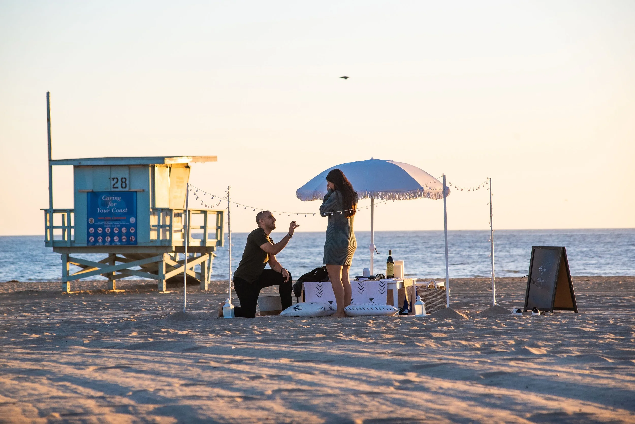 A man proposes marriage to a woman on a beach during sunset, with a small decorated setup including an umbrella, pillows, and a table with drinks, near a lifeguard tower.