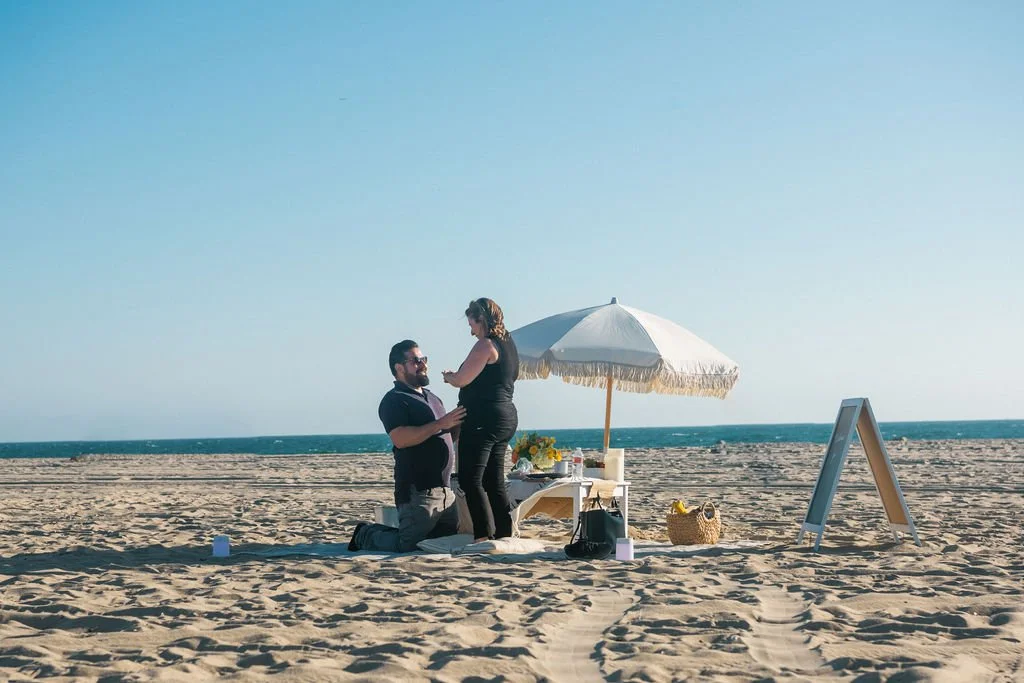 A couple having a picnic on a beach with a white umbrella, a table with food and drinks, a basket, and an easel sign.