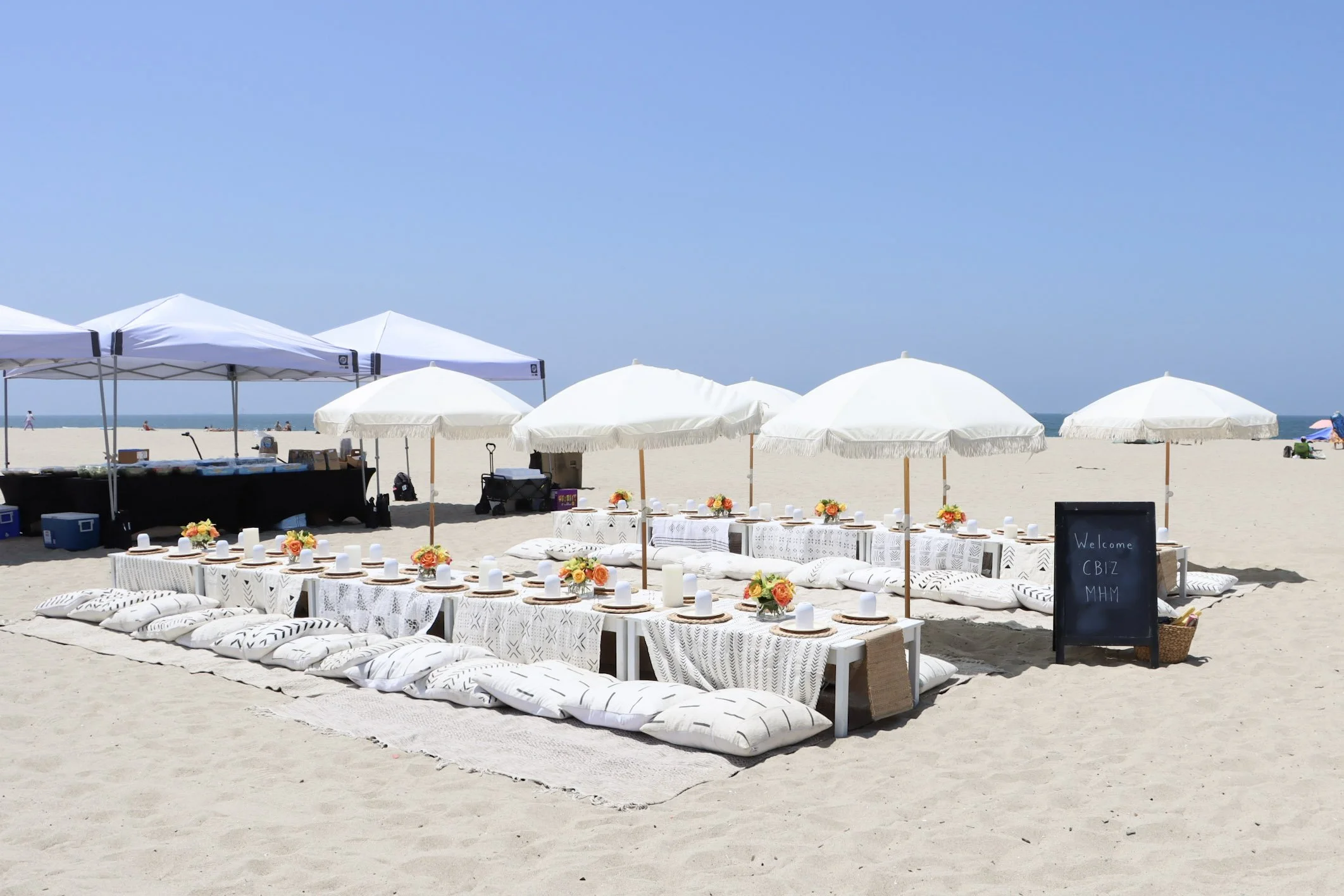 Beach dining setup with low white tables, white cushions, white umbrellas, floral centerpieces, and a black chalkboard sign, on sandy beach under a clear blue sky.