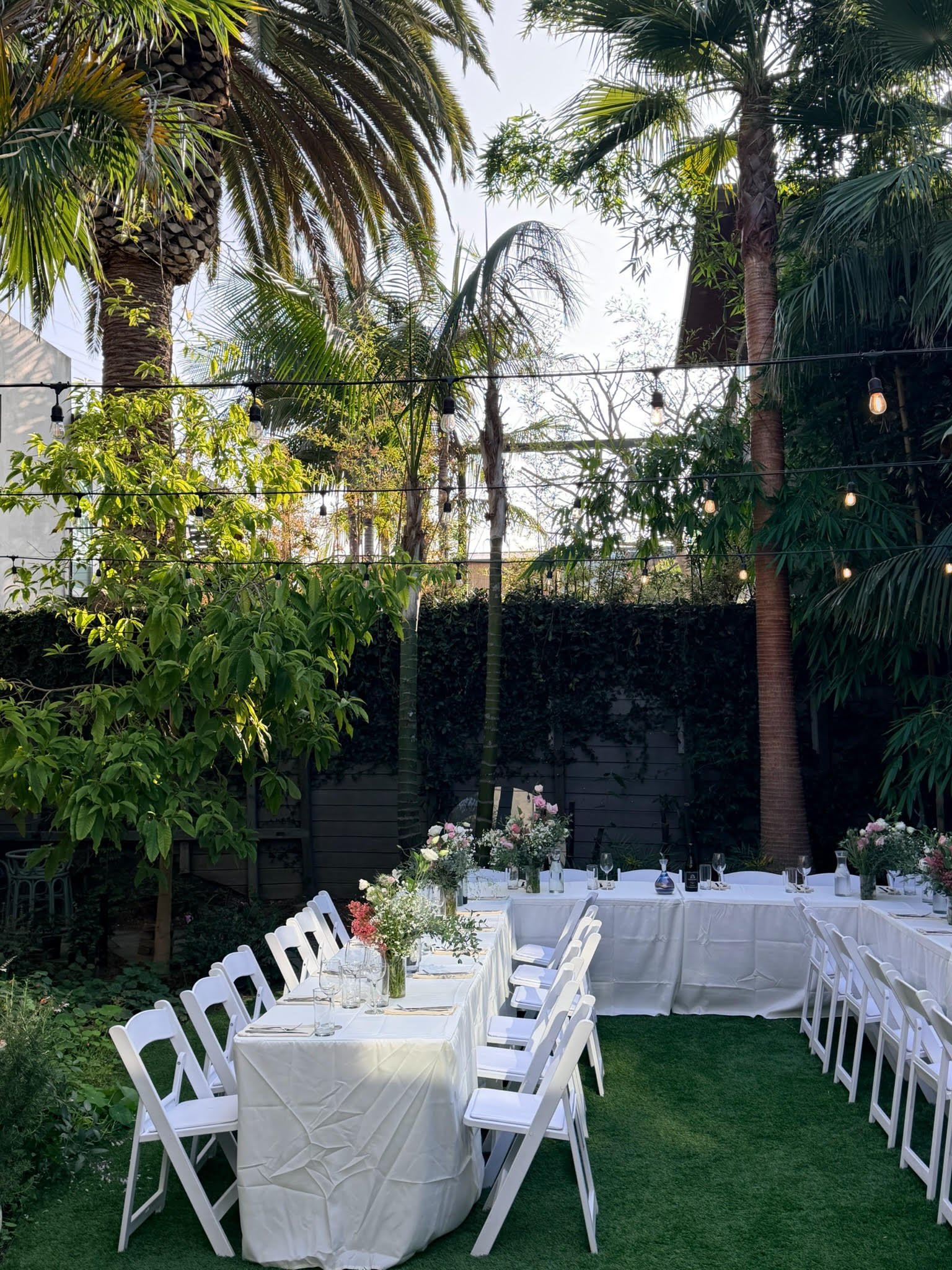 Outdoor dining area with a long table covered with a white tablecloth, set with glassware and floral centerpieces, surrounded by white chairs, under string lights, with lush green trees and plants in the background.