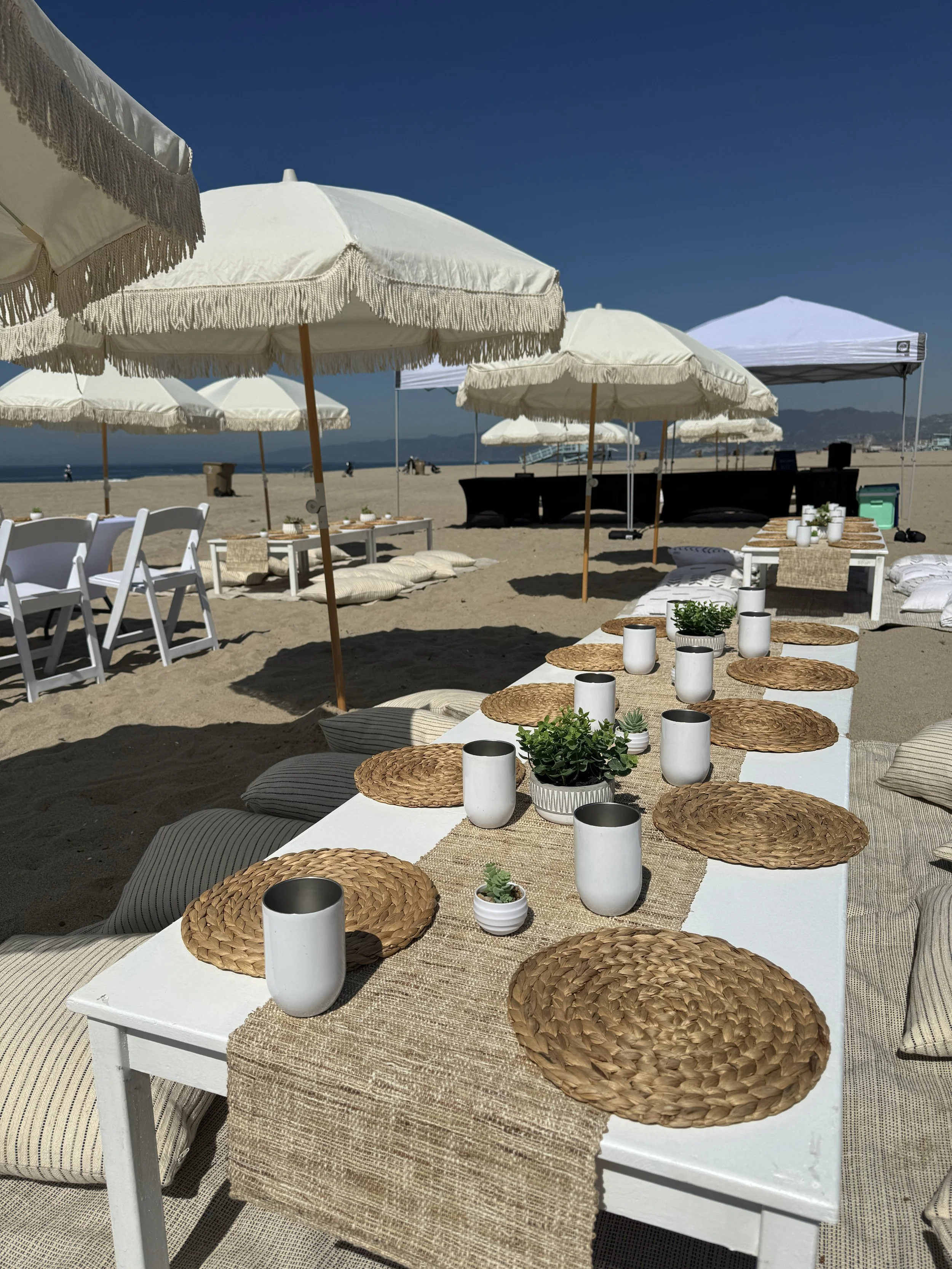 Beach setup with a long table, woven placemats, white cups, small potted plants, and pillows, under large white umbrellas on sandy beach, with ocean and mountains in background.