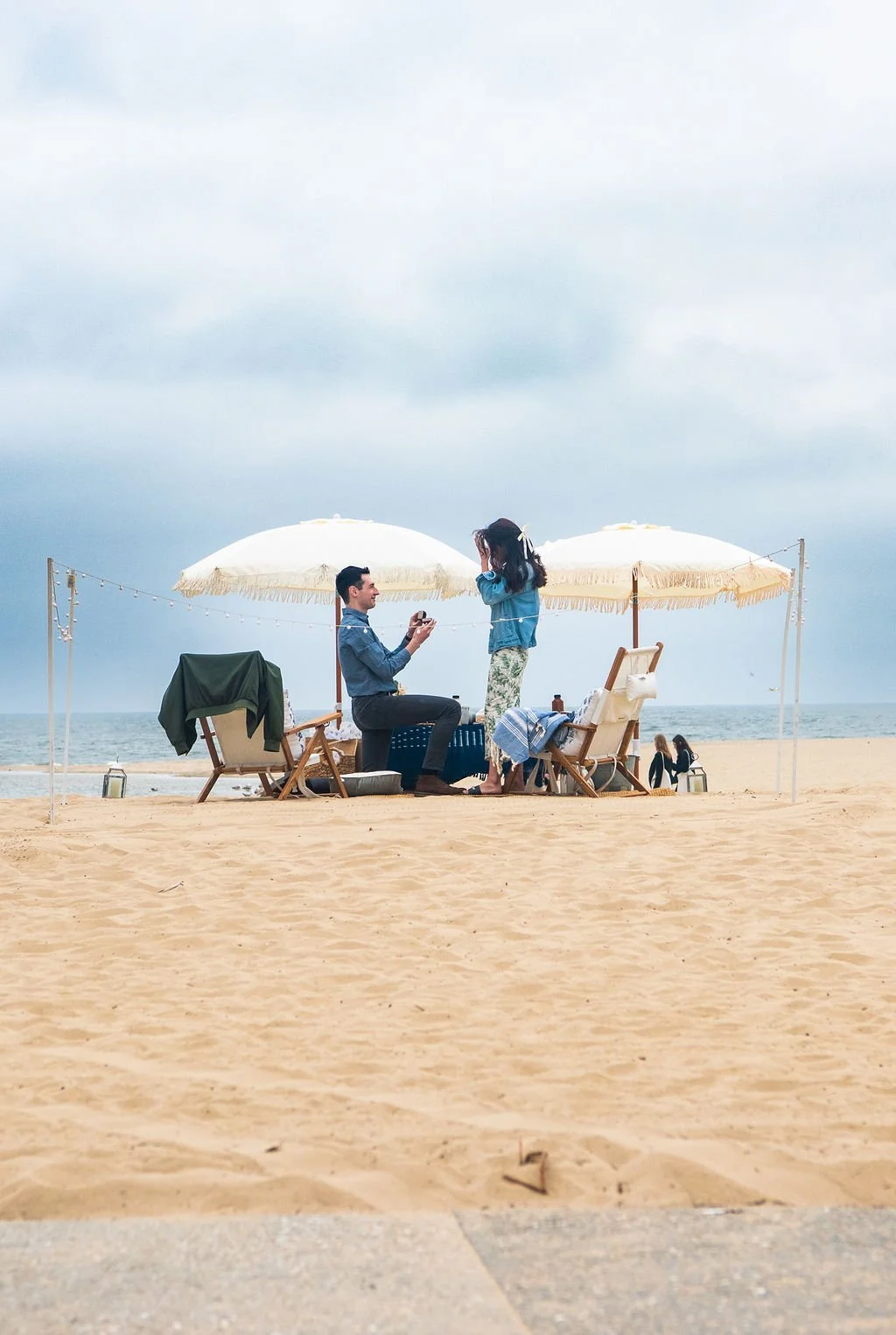 Couple on the beach under large white umbrellas, with chairs and a small table, engaged in a picnic or exchange, with sand and ocean in the background.