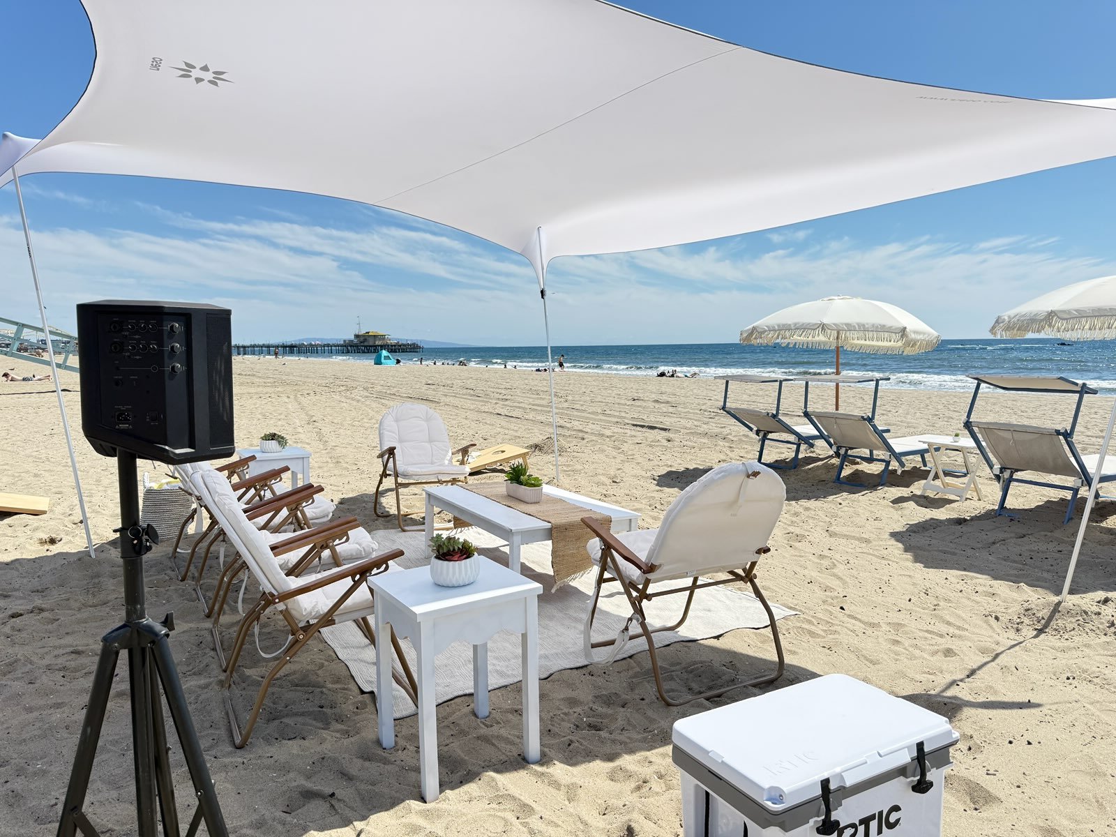 Beach scene with a shaded picnic area with a white table, striped chairs, coolers, and umbrellas. In the background, there are lounge chairs, umbrellas, and people on the sandy beach near the ocean, with a lifeguard tower and equipment visible on the right.