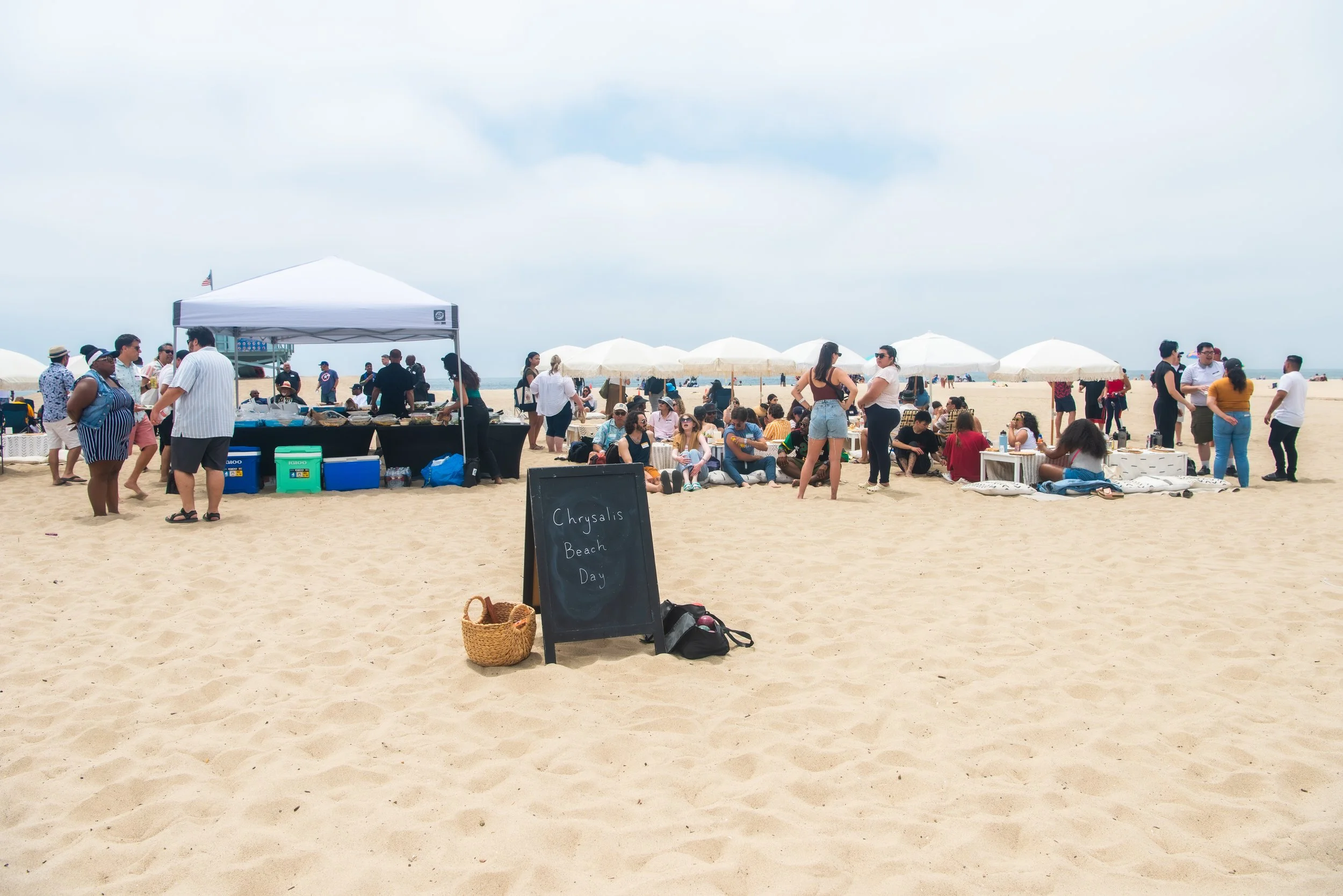 Crowd gathering at an outdoor beach event with tents, umbrellas, and people socializing on the sand during daytime.