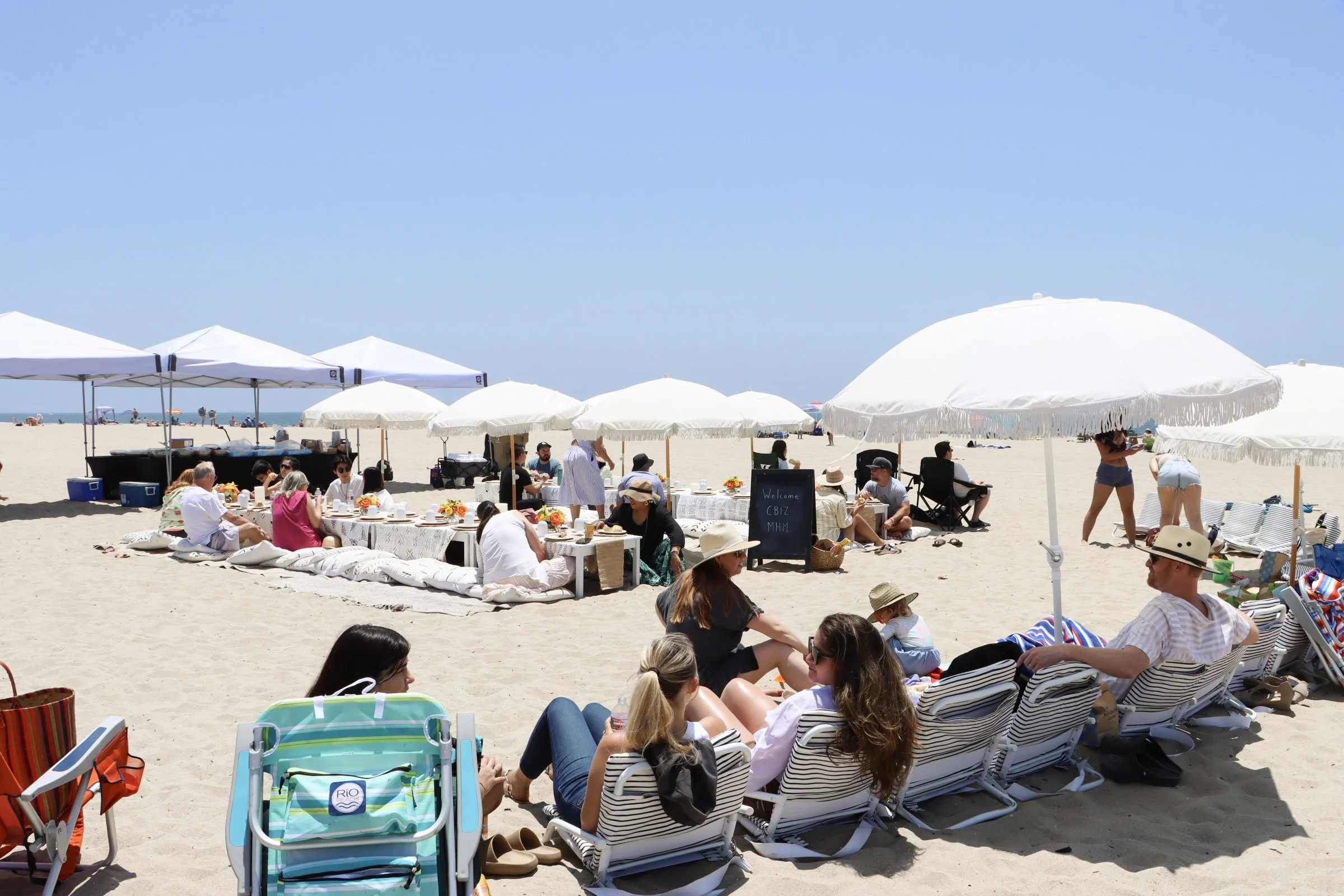 People enjoying a beach picnic under white umbrellas, sitting on cushions and beach chairs, with some eating at low tables, on a sandy beach under a clear blue sky.