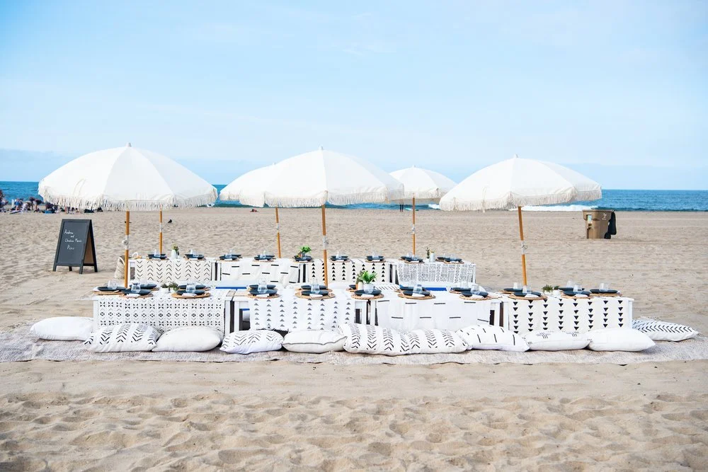Beach setup with white parasols, arranged table with black plates, cups, cutlery, and cushions on sand, overlooking ocean.