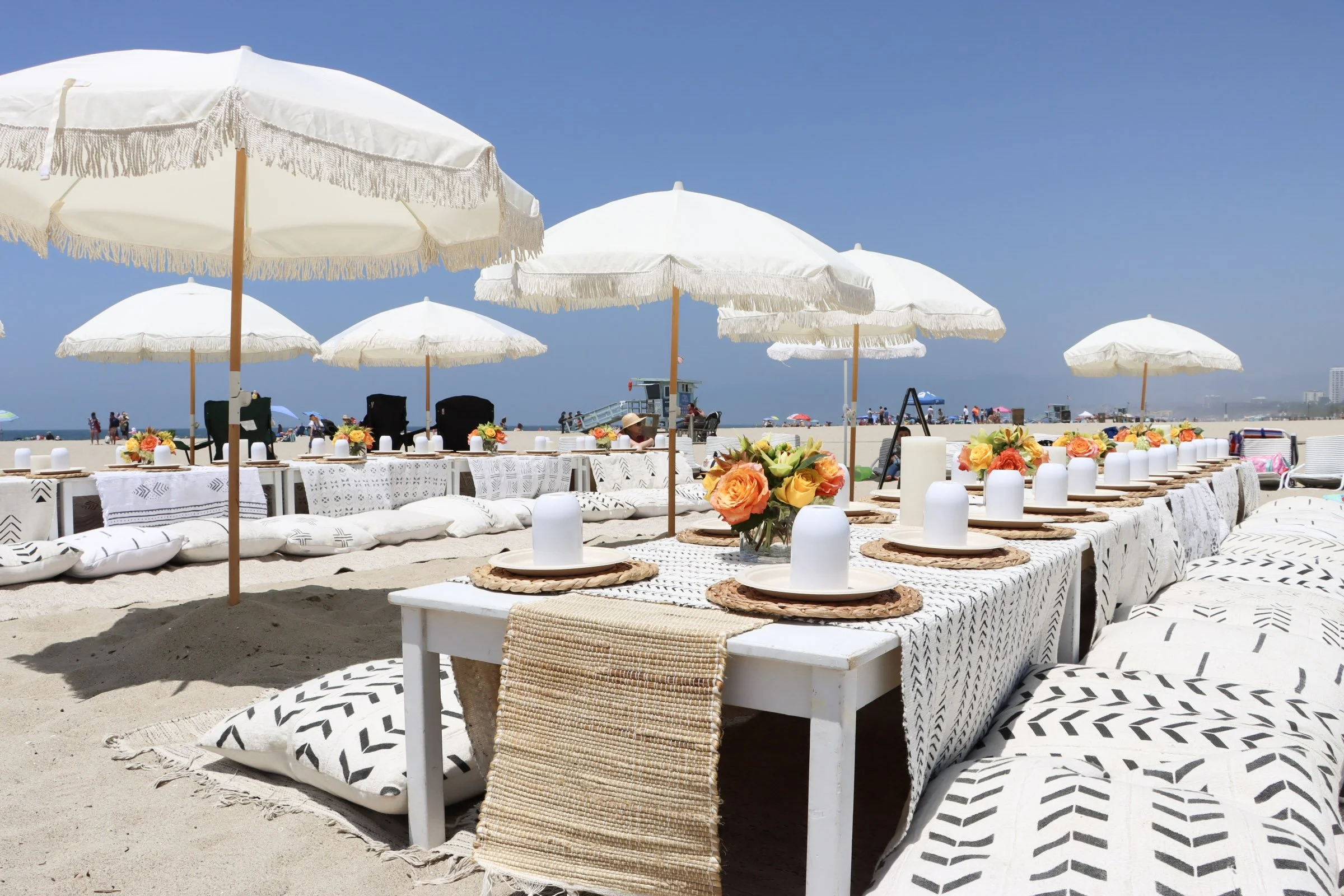 Beachside dining setup with white tables, black chairs, white parasols, floral centerpieces, and tableware on sandy beach under clear blue sky.