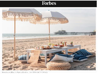 Beach setup with a table, food, and drinks under umbrellas on the sand near the ocean, with a pier in the background.