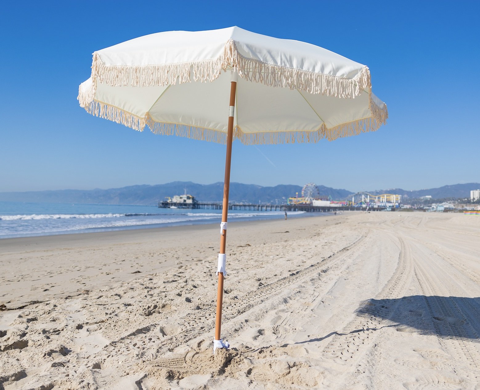A white beach umbrella with fringes on a sandy beach with a pier and amusement park in the background, under a clear blue sky.