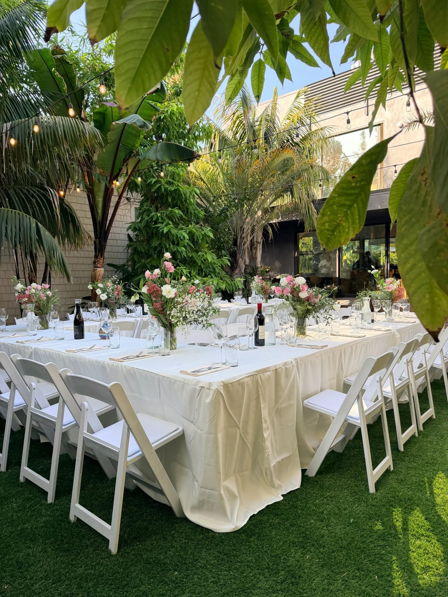 Outdoor dining setup with a long white table covered with a white tablecloth, decorated with pink and white floral centerpieces. White chairs surround the table. The setting is in a lush, green garden with tropical plants and string lights overhead.