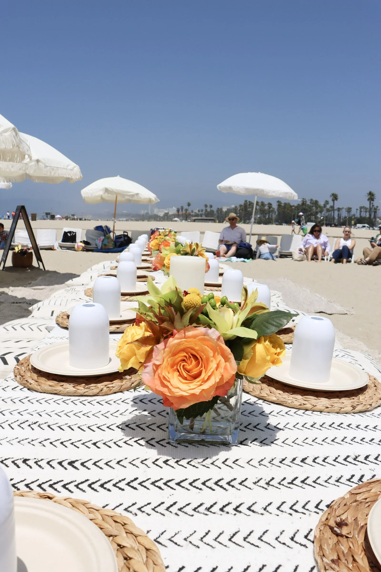 A long outdoor dining table set on a beach with floral centerpieces, plates, and glasses, with umbrellas, beach chairs, and people relaxing in the background under a clear blue sky.