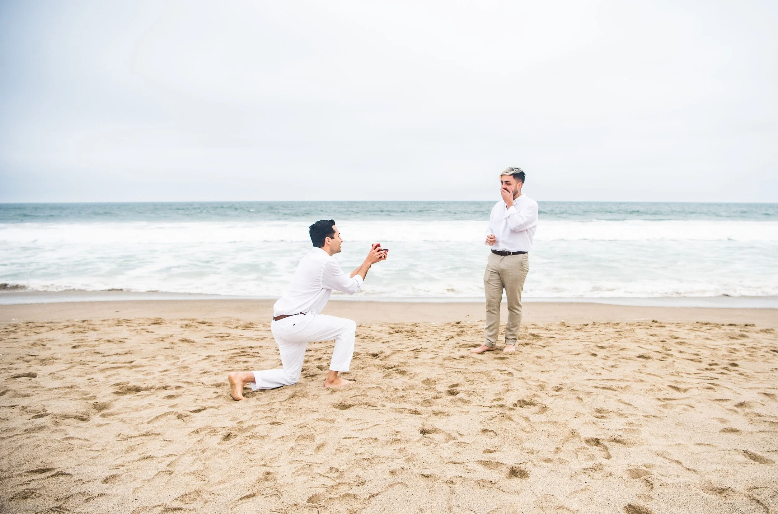 A man proposing marriage on a beach with another man kneeling and holding a ring, both in white shirts and beige pants, with the ocean in the background.
