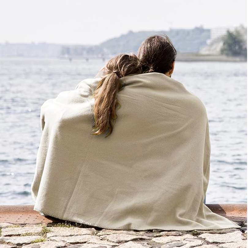 A person and a child sitting by the water wrapped in a beige blanket, with city buildings and hills in the background.