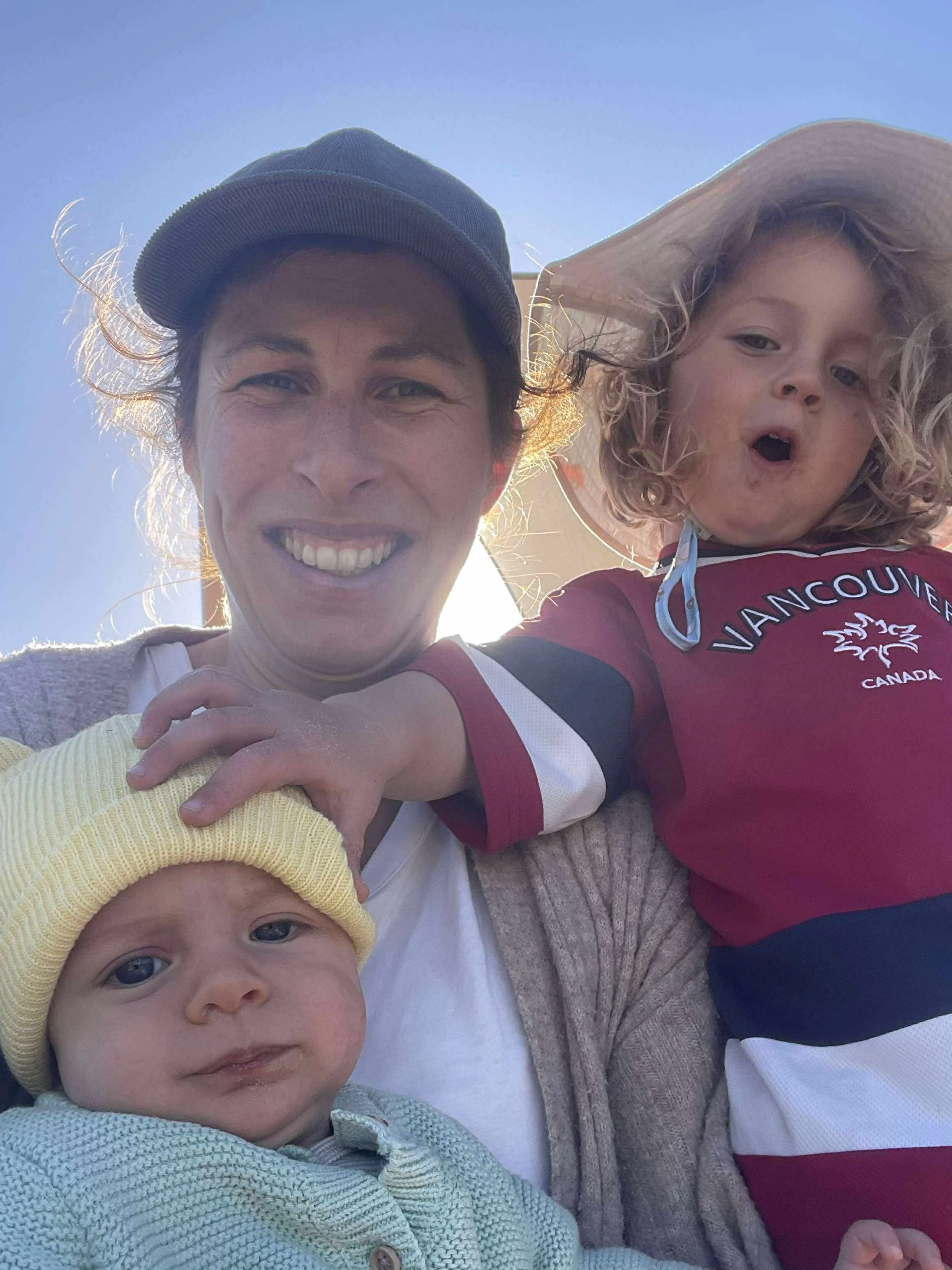 A smiling woman with two young children outdoors, one child wearing a yellow beanie and the other in a red Vancouver Canada shirt and sun hat, with blue sky in the background.