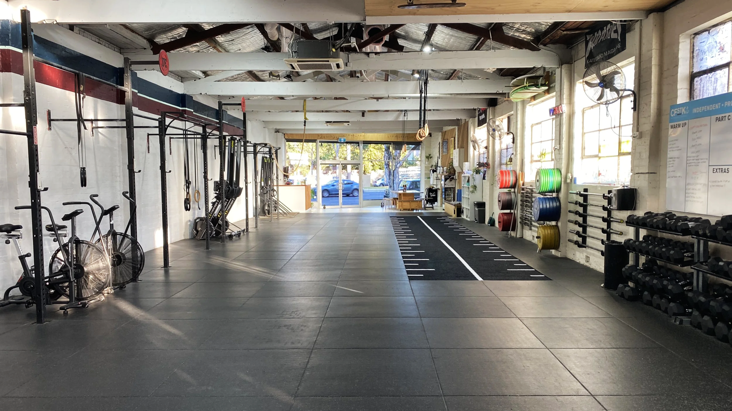 Empty gym with workout equipment, weights, and a treadmill, sunlight coming through large windows.