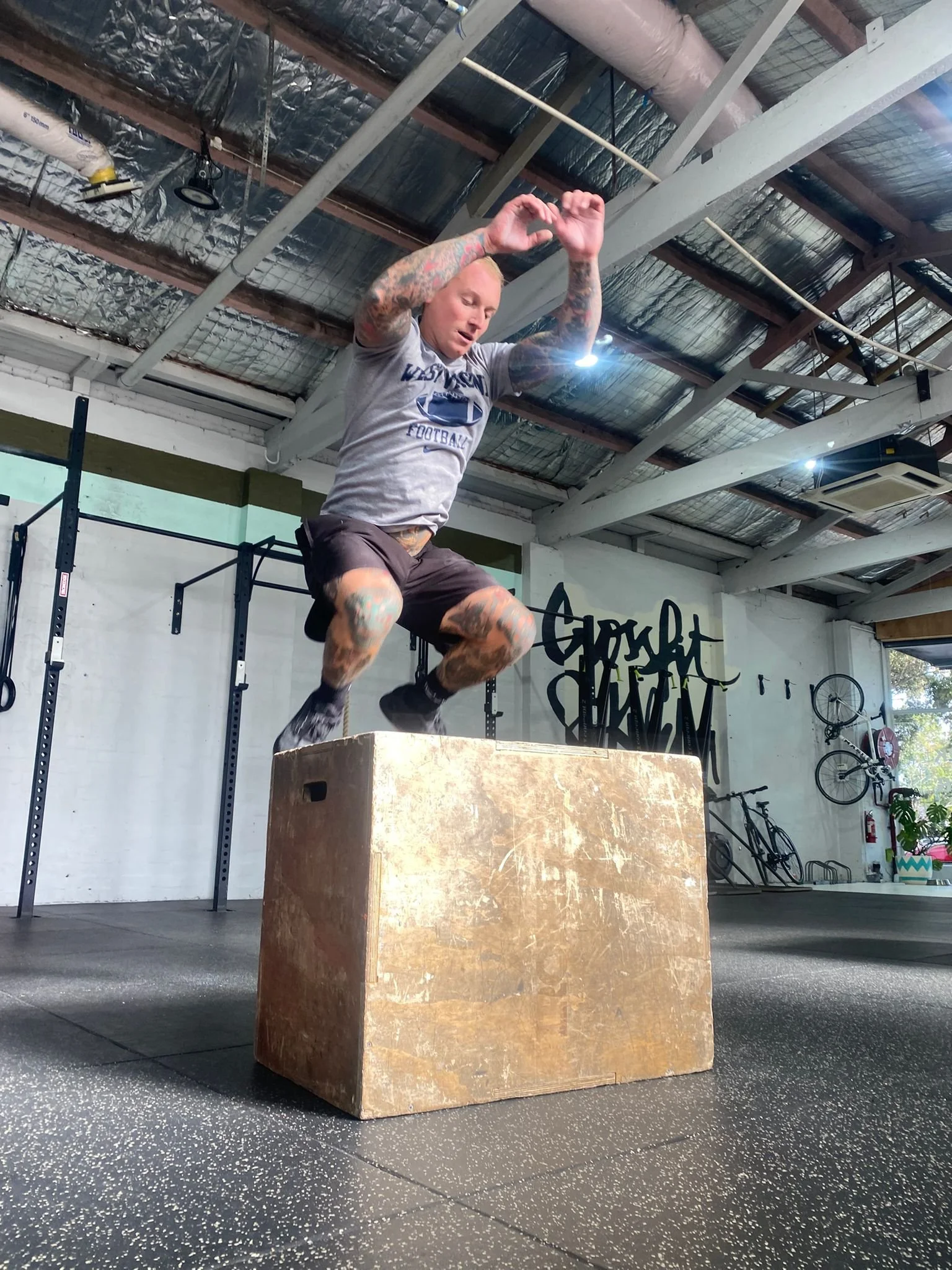 A man with tattoos jumping onto a large wooden box in a gym.