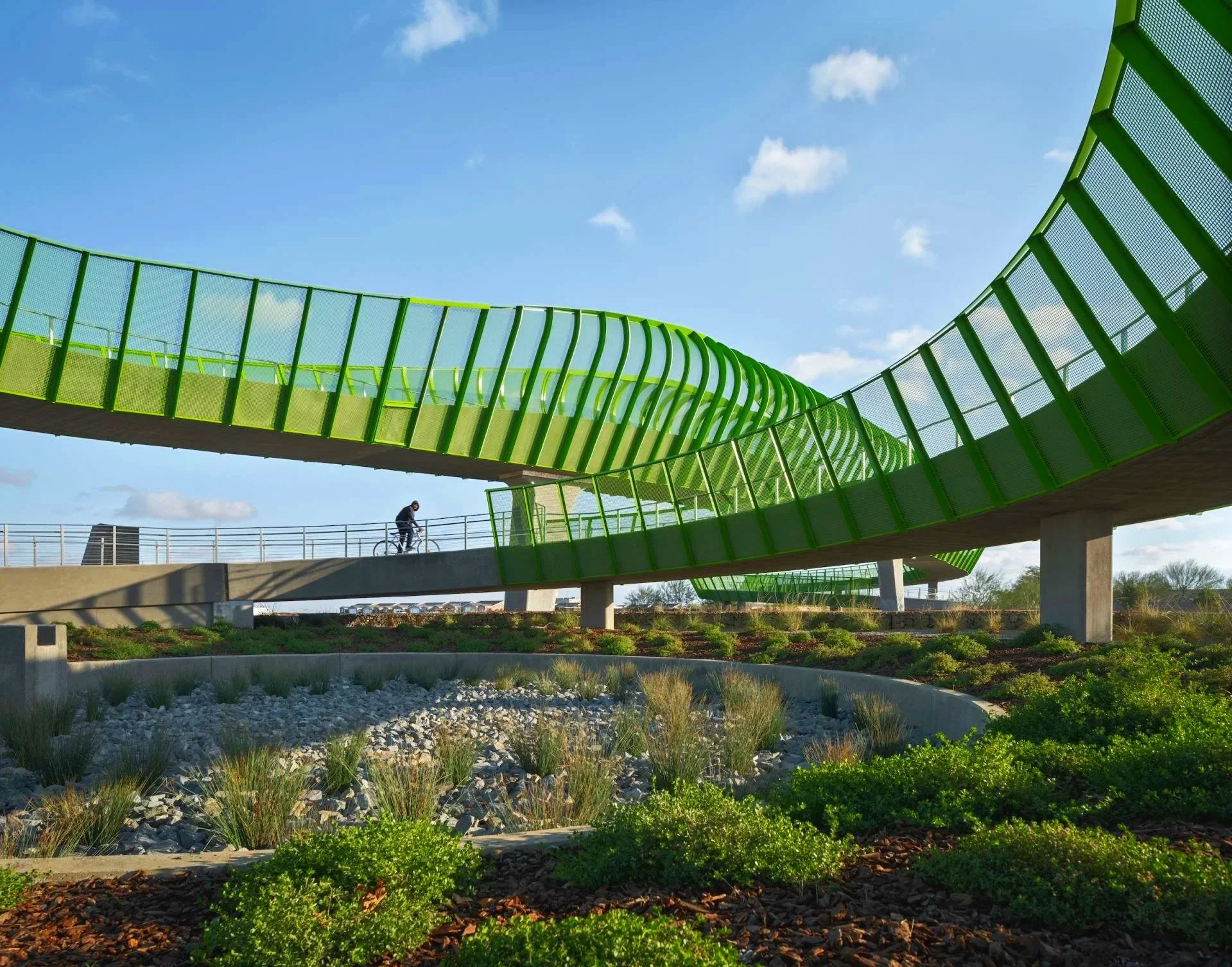 A modern, curved pedestrian bridge painted bright green with a metal mesh railing, spanning over a dry landscaped area with rocks and native plants, under a partly cloudy blue sky.