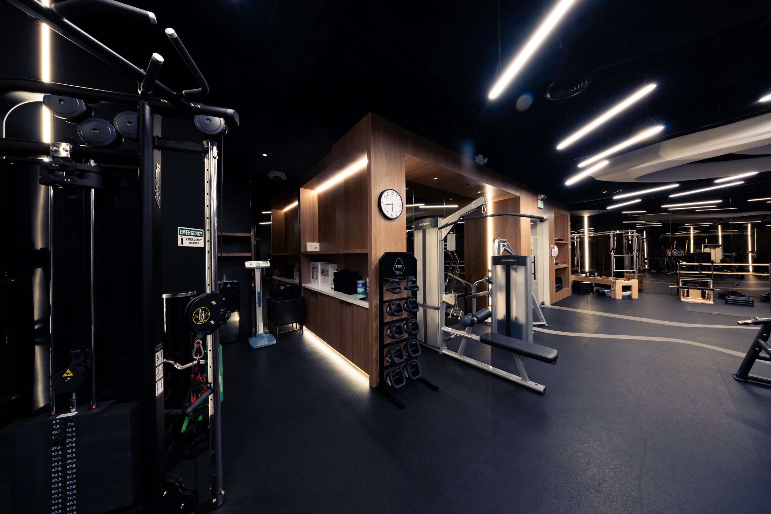 Interior of a modern gym at Blair House Condominium, Los Angeles with various workout machines, weights, and equipment illuminated by ceiling lights and a wooden partition.