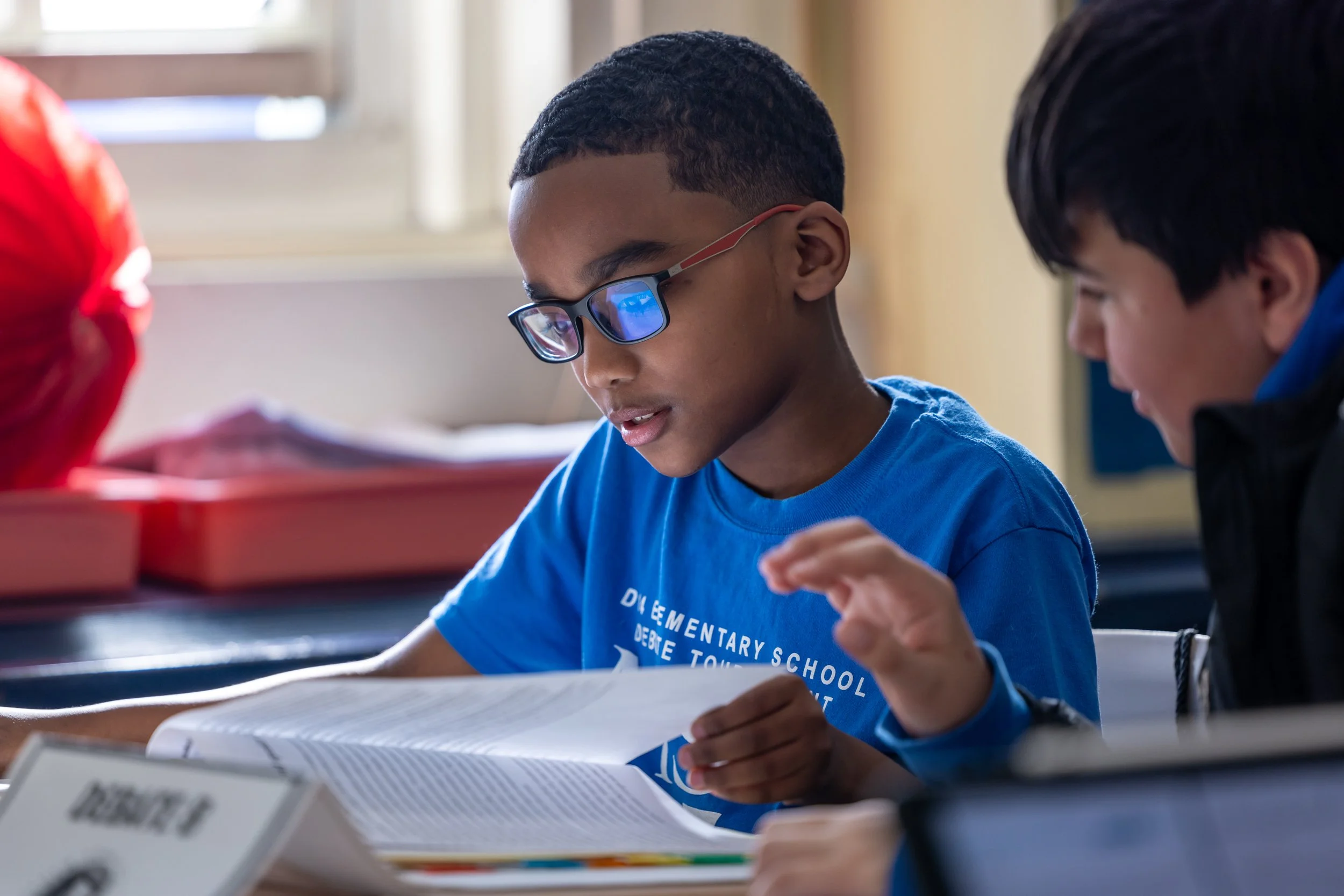 A young student with glasses reading a textbook during class with a classmate.
