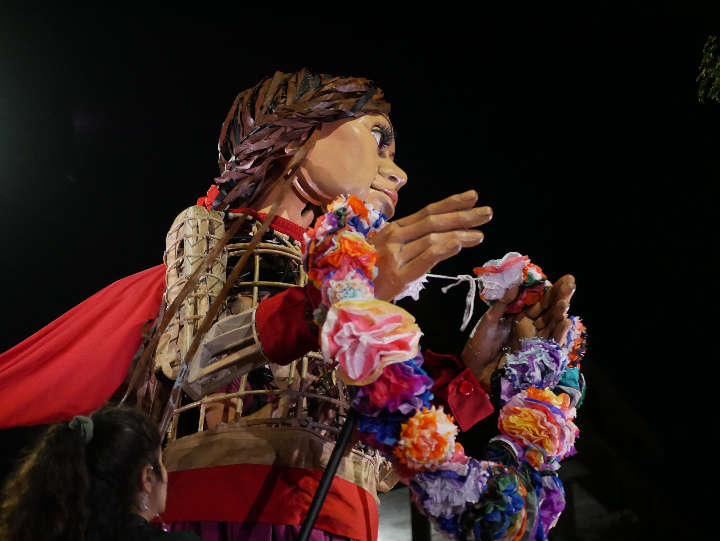 Little Amal holds up the lei of flowers