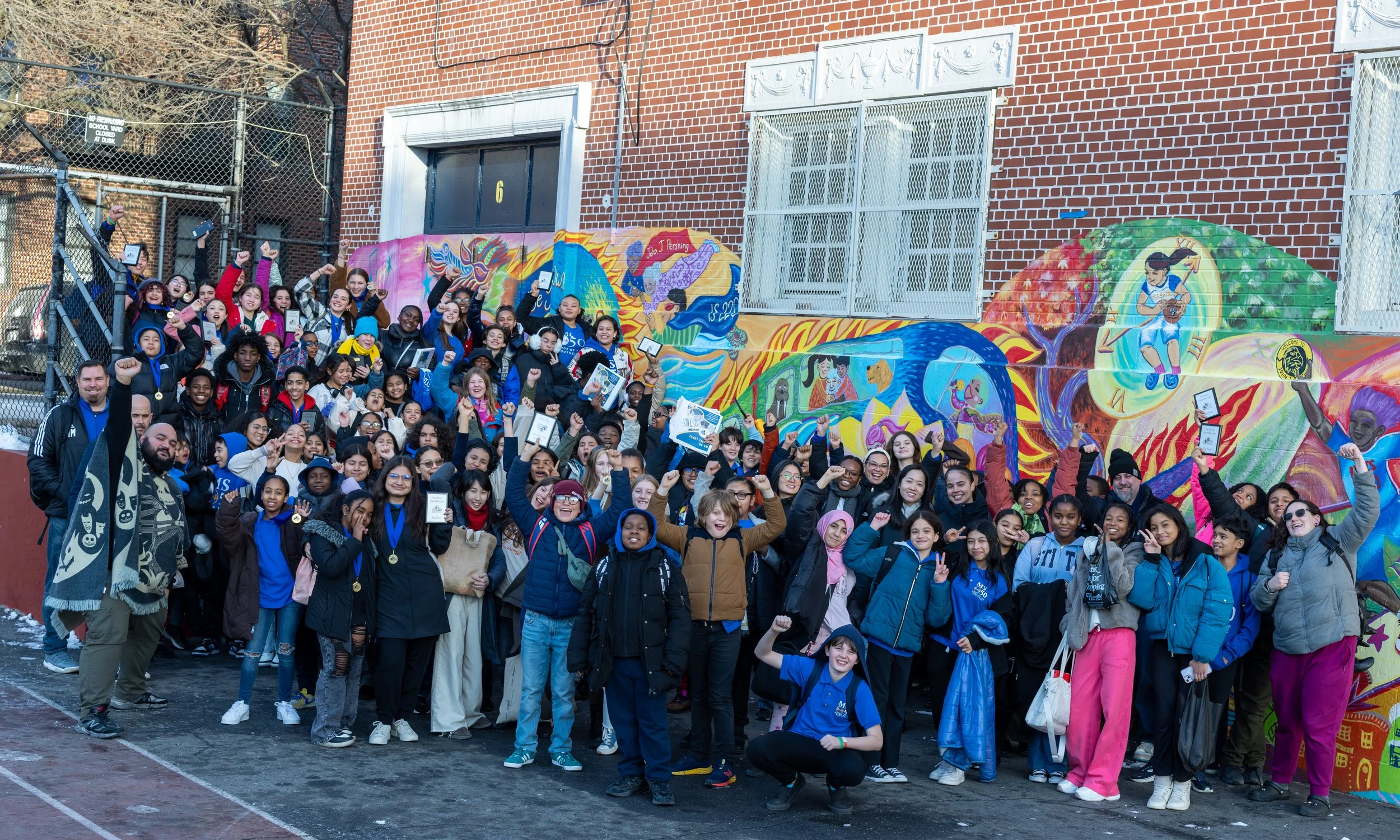 Group of students and teachers celebrating outdoors in front of a colorful mural, some holding awards or medals.
