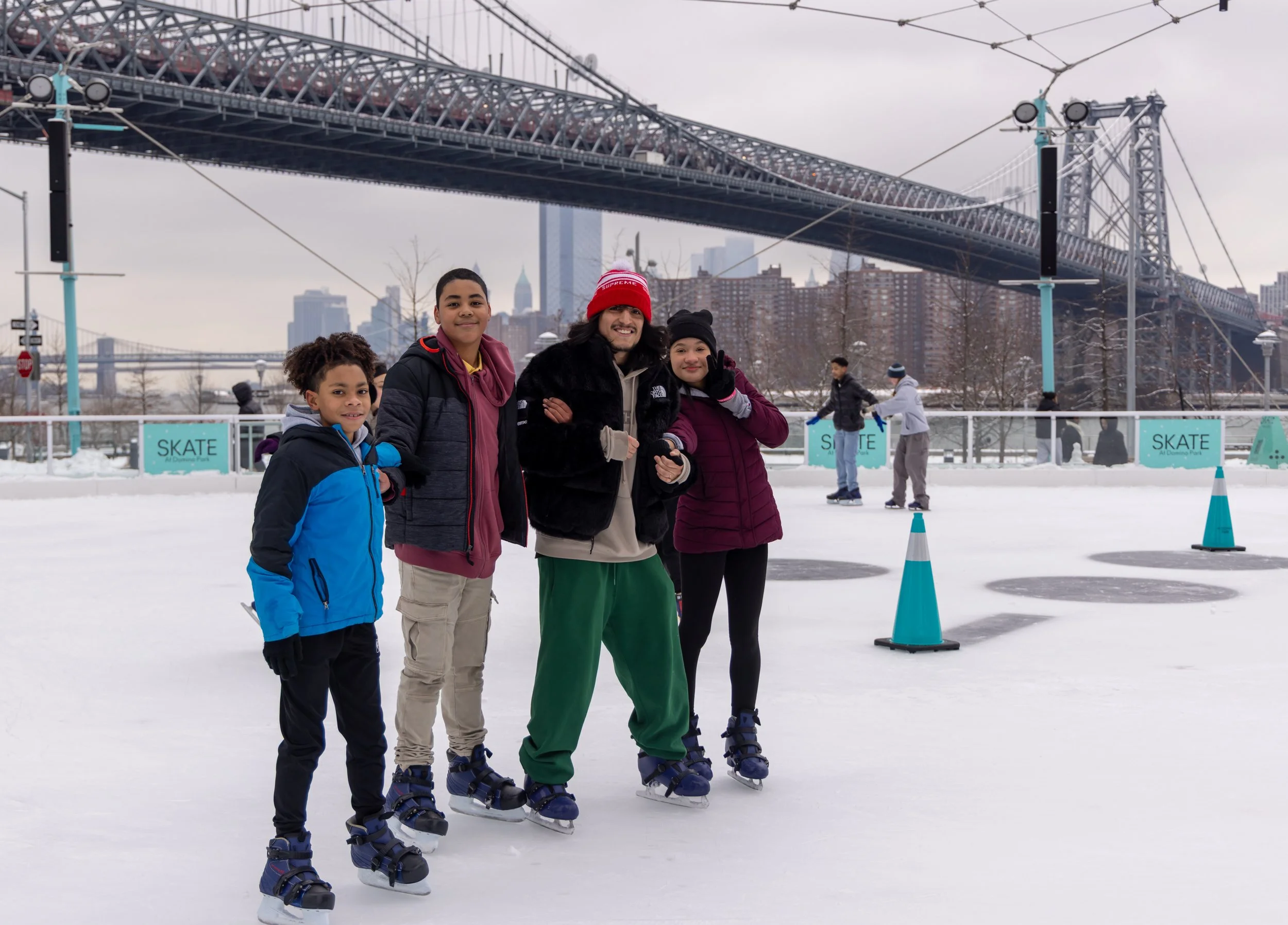 A group of four people ice skating outdoors on an ice rink with city skyscrapers in the background and a large bridge overhead. They are wearing winter clothing and skates.