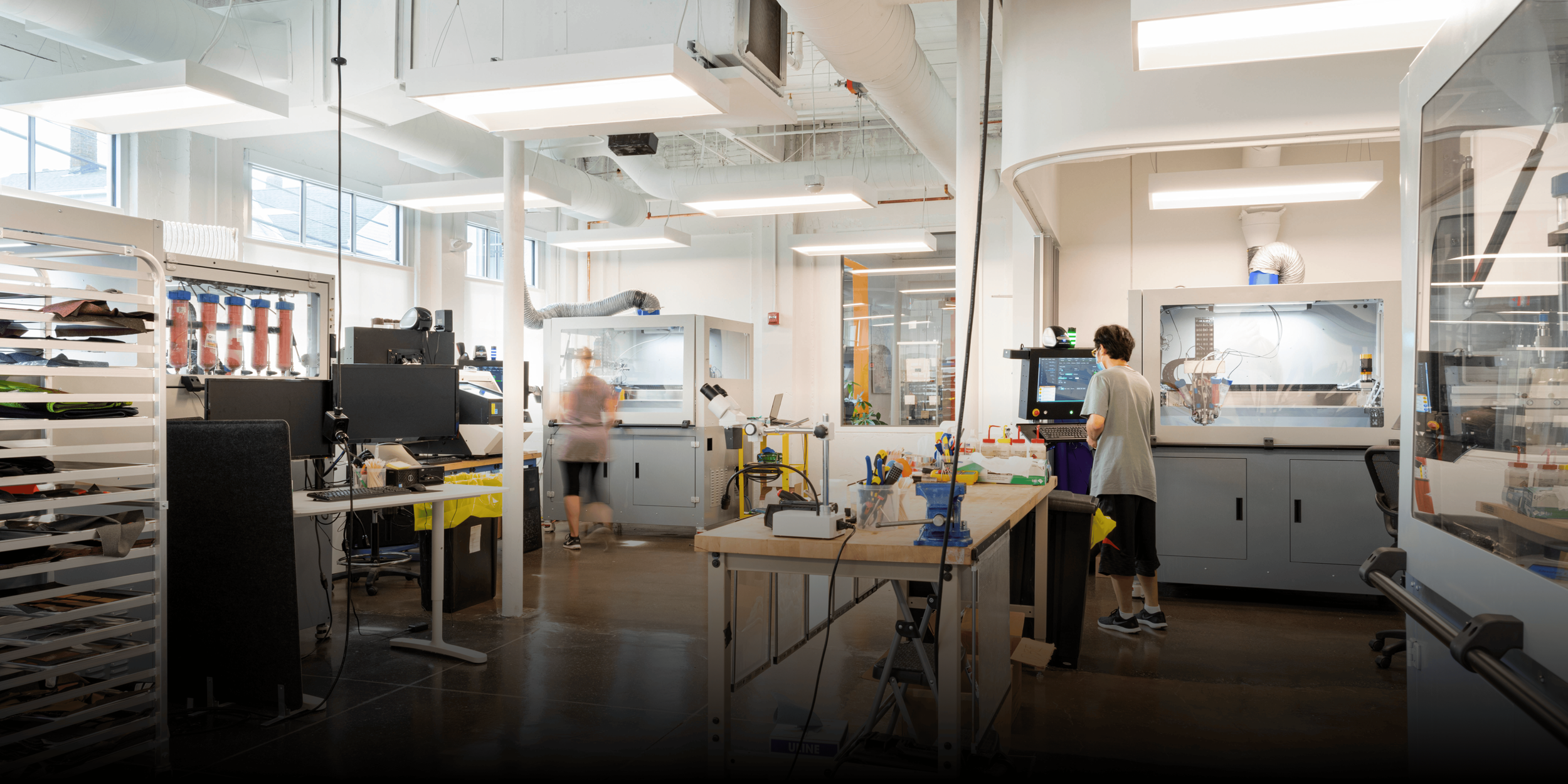 A modern industrial workspace with multiple workers operating machines and computer systems, surrounded by equipment, tools, and storage units.