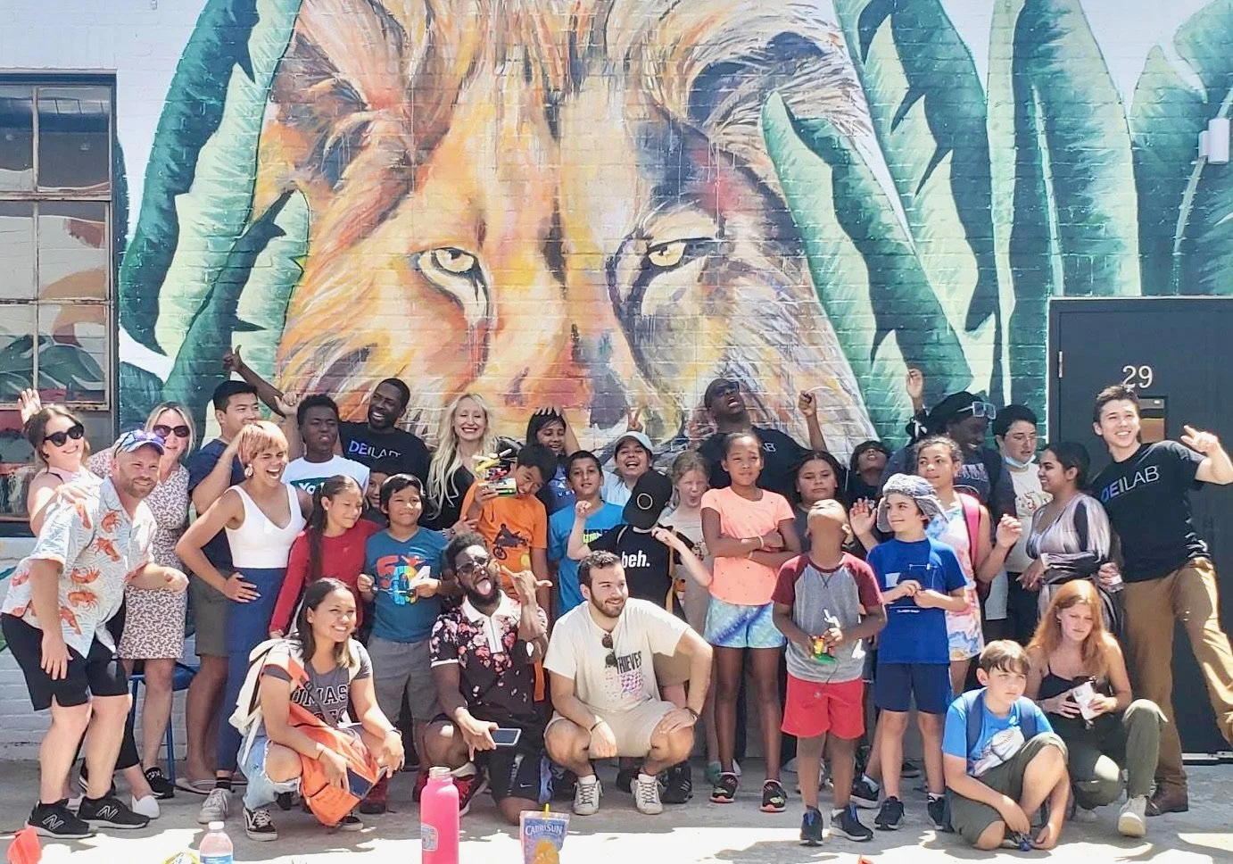 Group of children and adults posing for a photo in front of a large colorful mural of a lion on a brick wall.