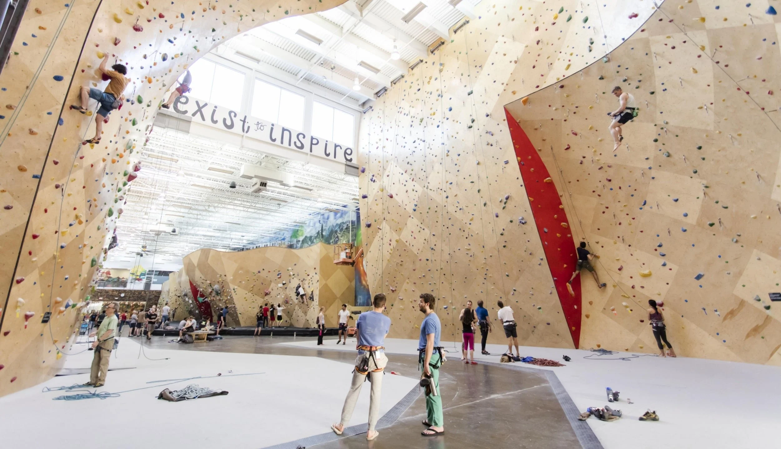 Indoor rock climbing gym with climbers on various routes on beige and red climbing walls. There are people belaying and preparing gear, with climbing shoes and ropes on the floor. A large window lets in natural light, and a sign in the background rea