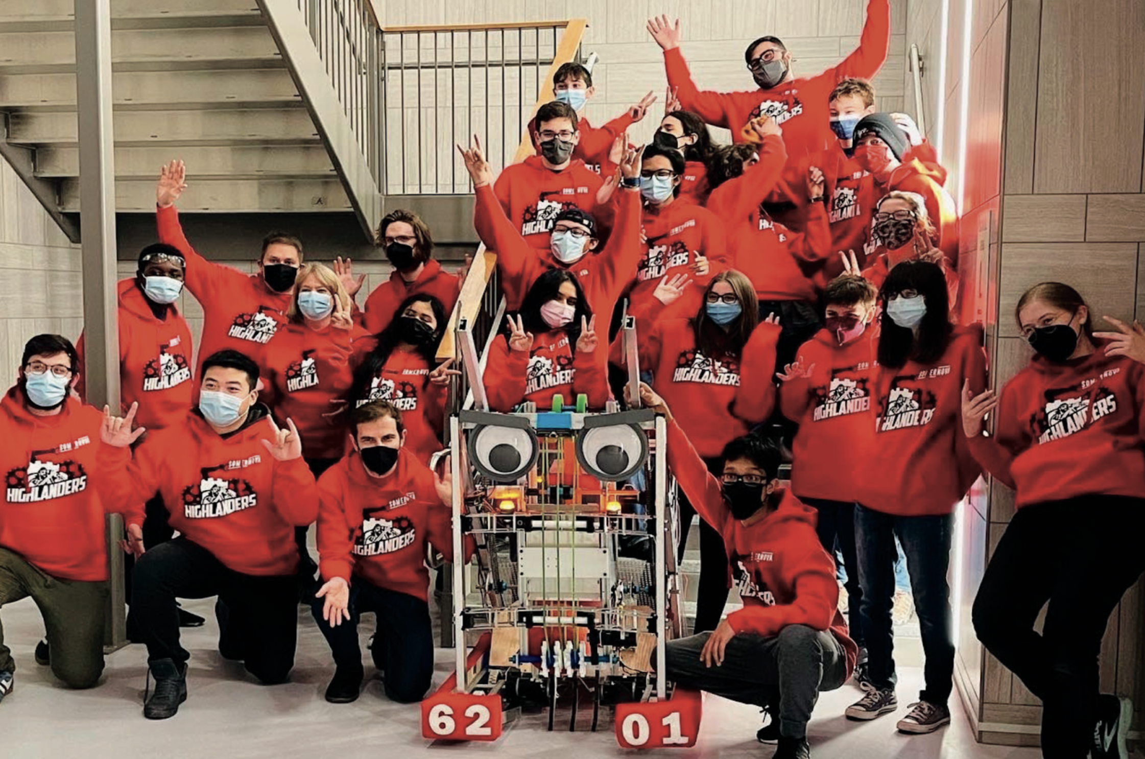 A large group of people wearing red hoodies and face masks, gathered around a robot in an indoor setting, celebrating or posing for a photo.
