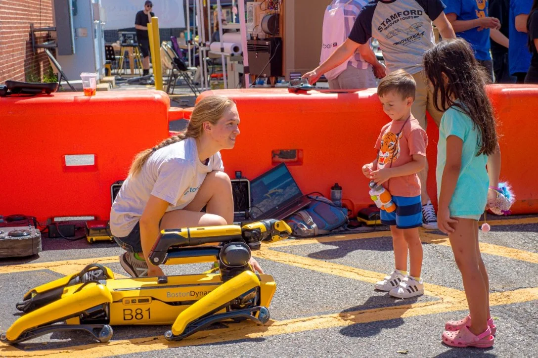 A woman is explaining a yellow Boston Dynamics robotic robot to a young boy and girl in a parking lot, with other people and equipment in the background.