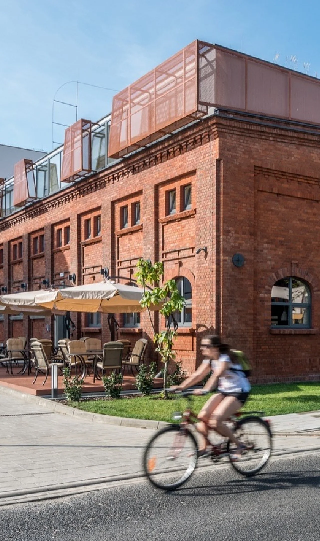A person riding a bicycle on the sidewalk in front of a red brick building with outdoor seating and modern orange balconies on the upper floor, under a clear blue sky.
