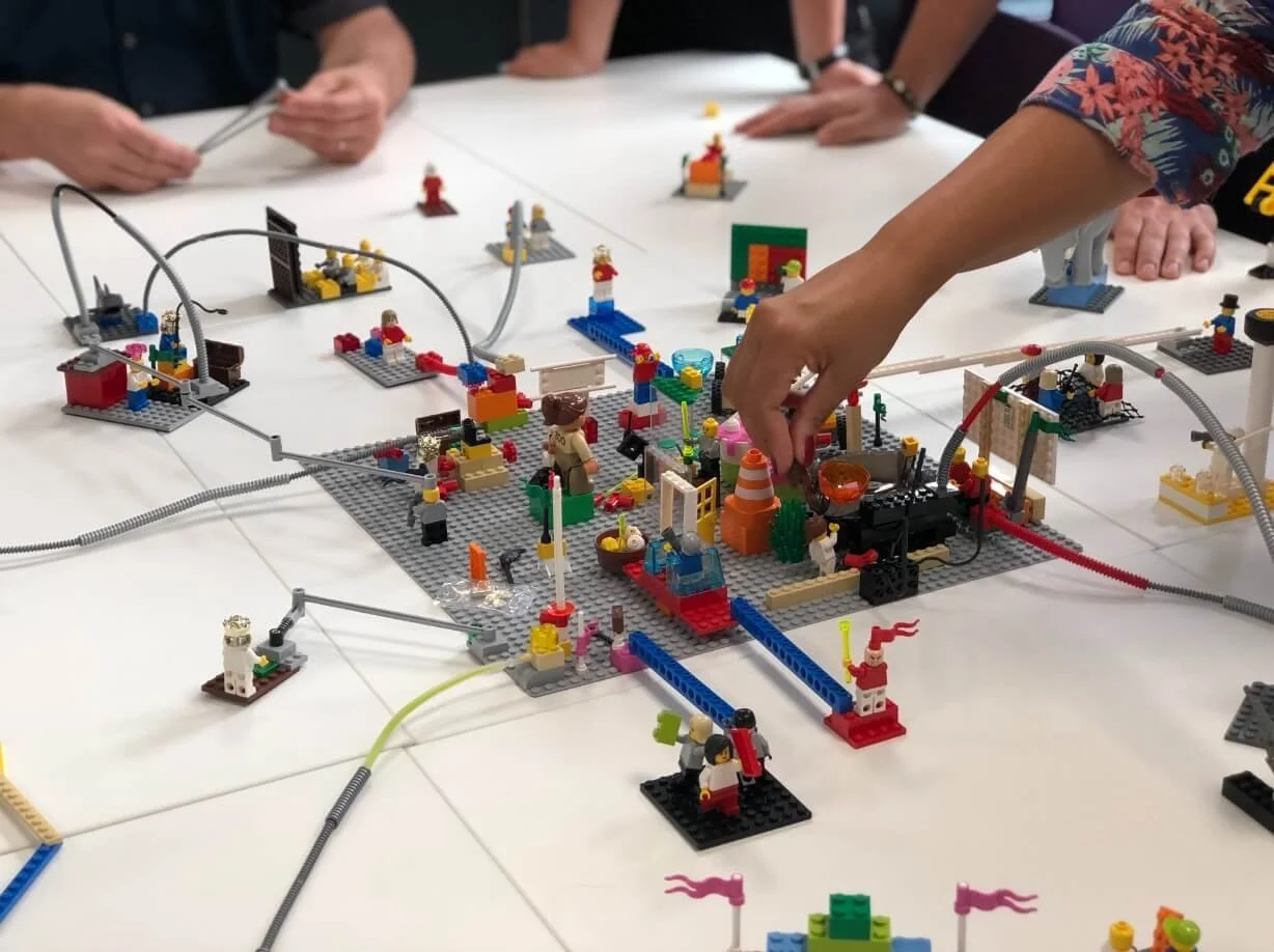 People playing with LEGO building blocks on a white table, creating a detailed construction scene.