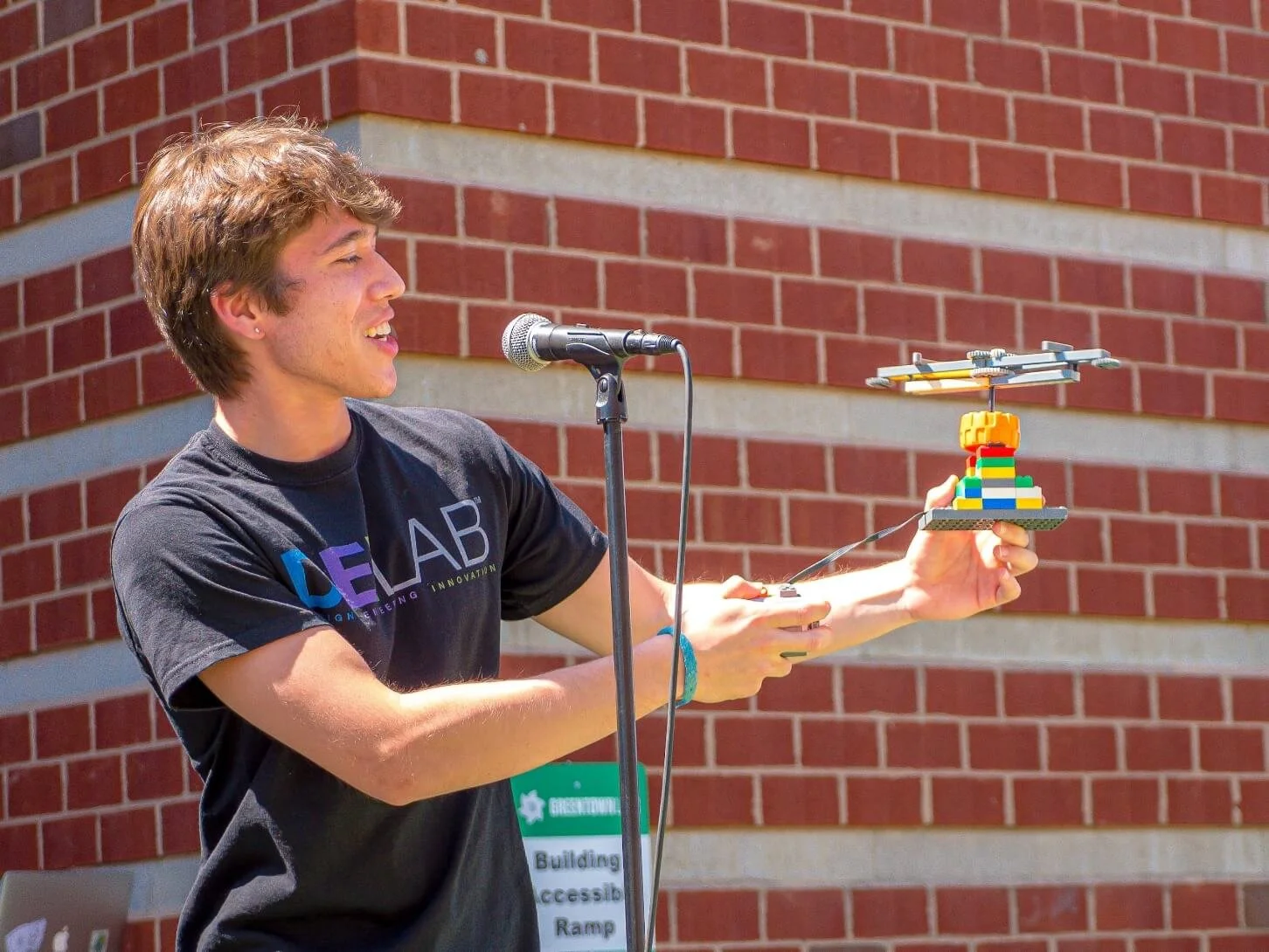 A young man with brown hair speaking into a microphone outdoors, holding a LEGO helicopter built on a LEGO base, in front of a brick building.