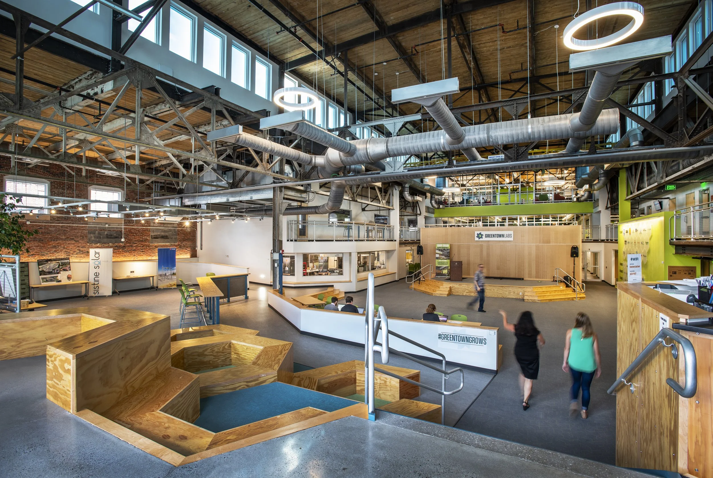 Interior of a modern, industrial-style building with exposed ductwork, large windows, and a high wooden ceiling. There are wooden steps, seating areas, and people walking and sitting inside, with a sign that reads 'GREENTOWN GROWS' on a wooden wall.