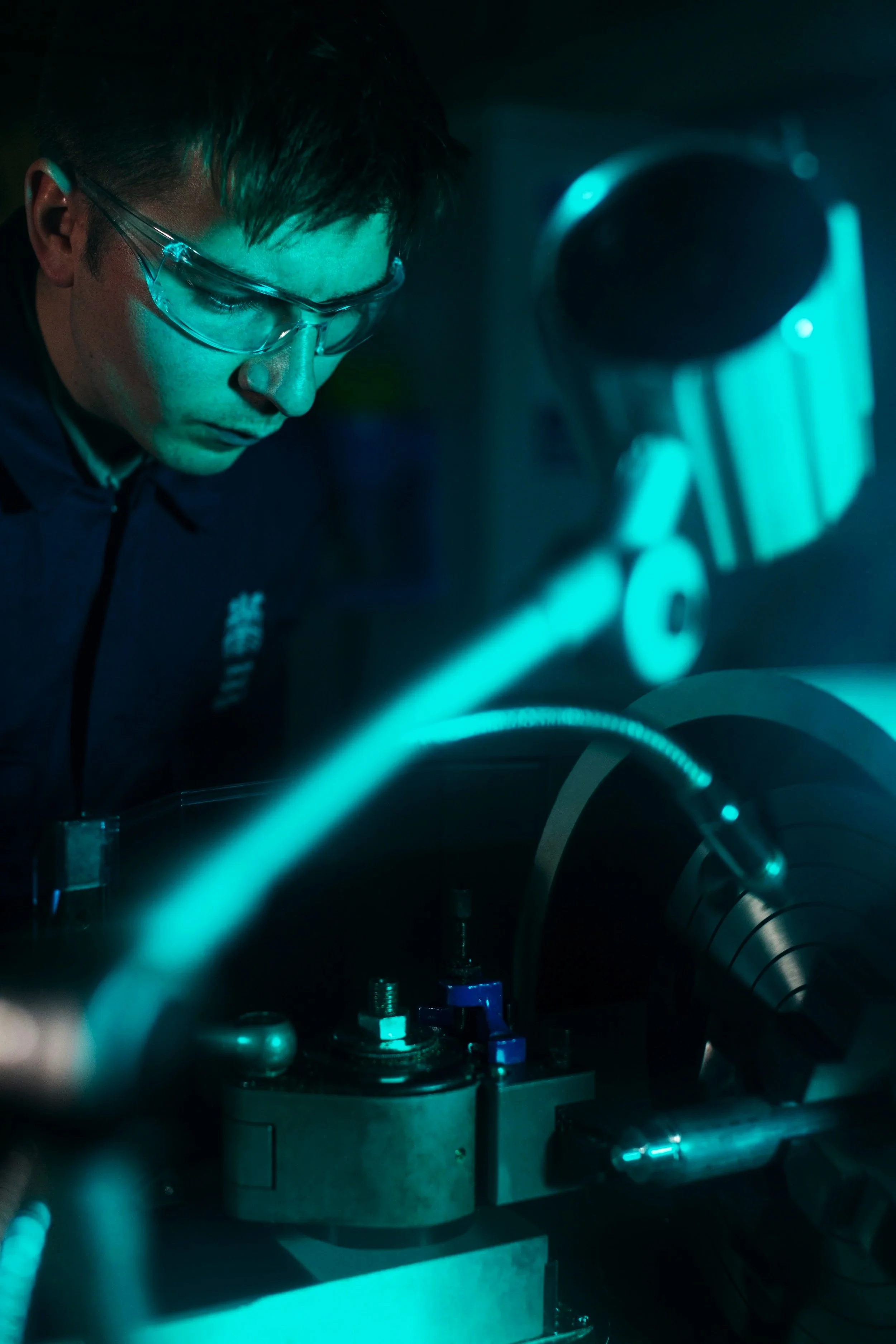A scientist wearing safety glasses analyzing a sample through a microscope in a laboratory with dark lighting and blue hues.