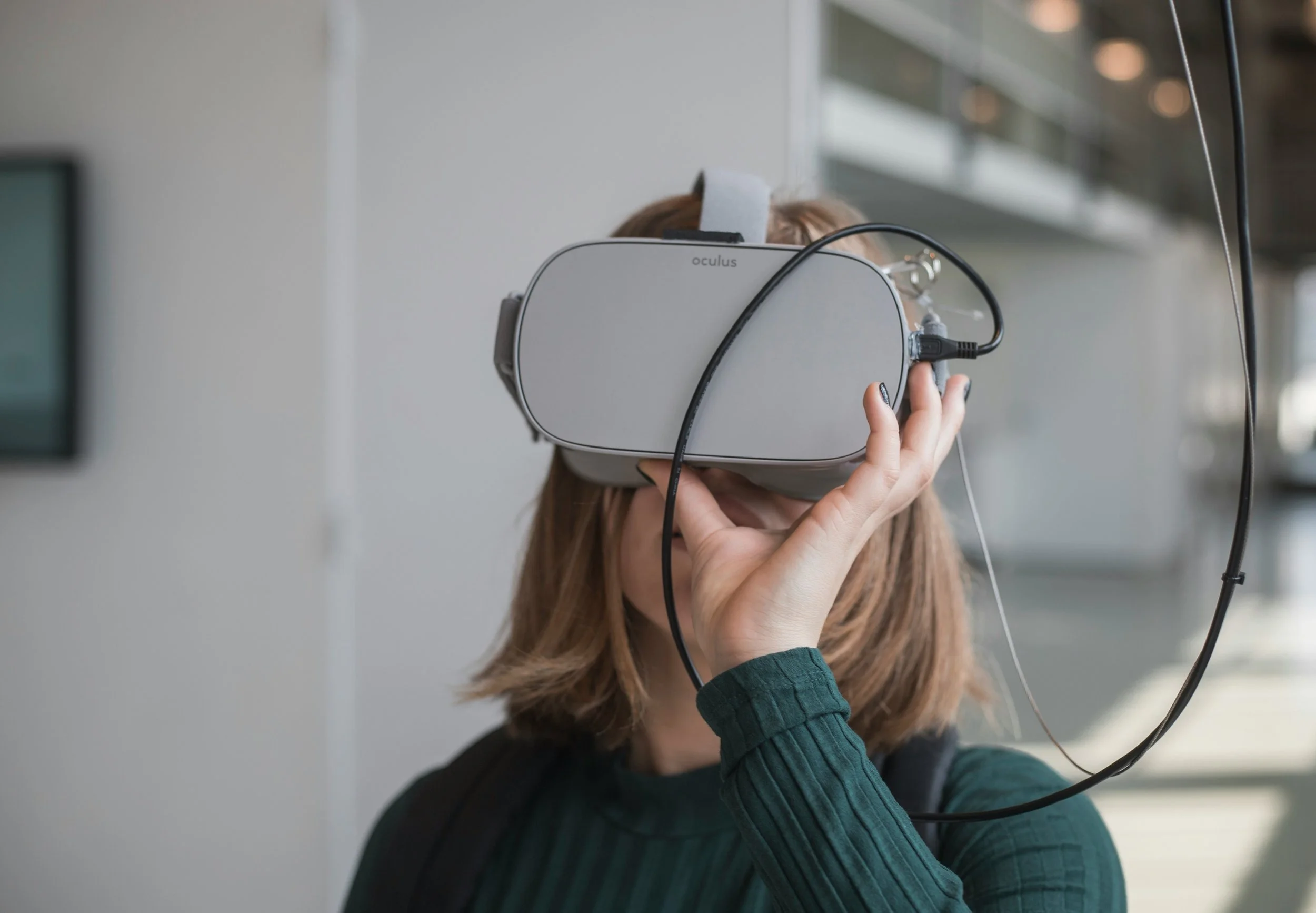 A person wearing a virtual reality headset with the brand 'Oculus' is holding a controller in one hand, engaged in a virtual reality experience. The person is indoors, near a window with a blurred cityscape in the background.