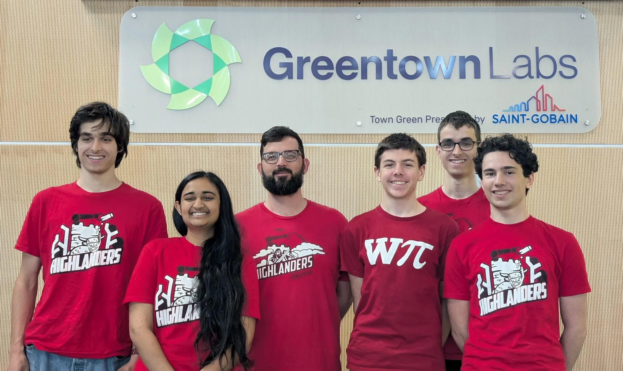 Group of six young people standing in front of a gray wall with a Greentown Labs sign, all wearing red Highlanders T-shirts and smiling.